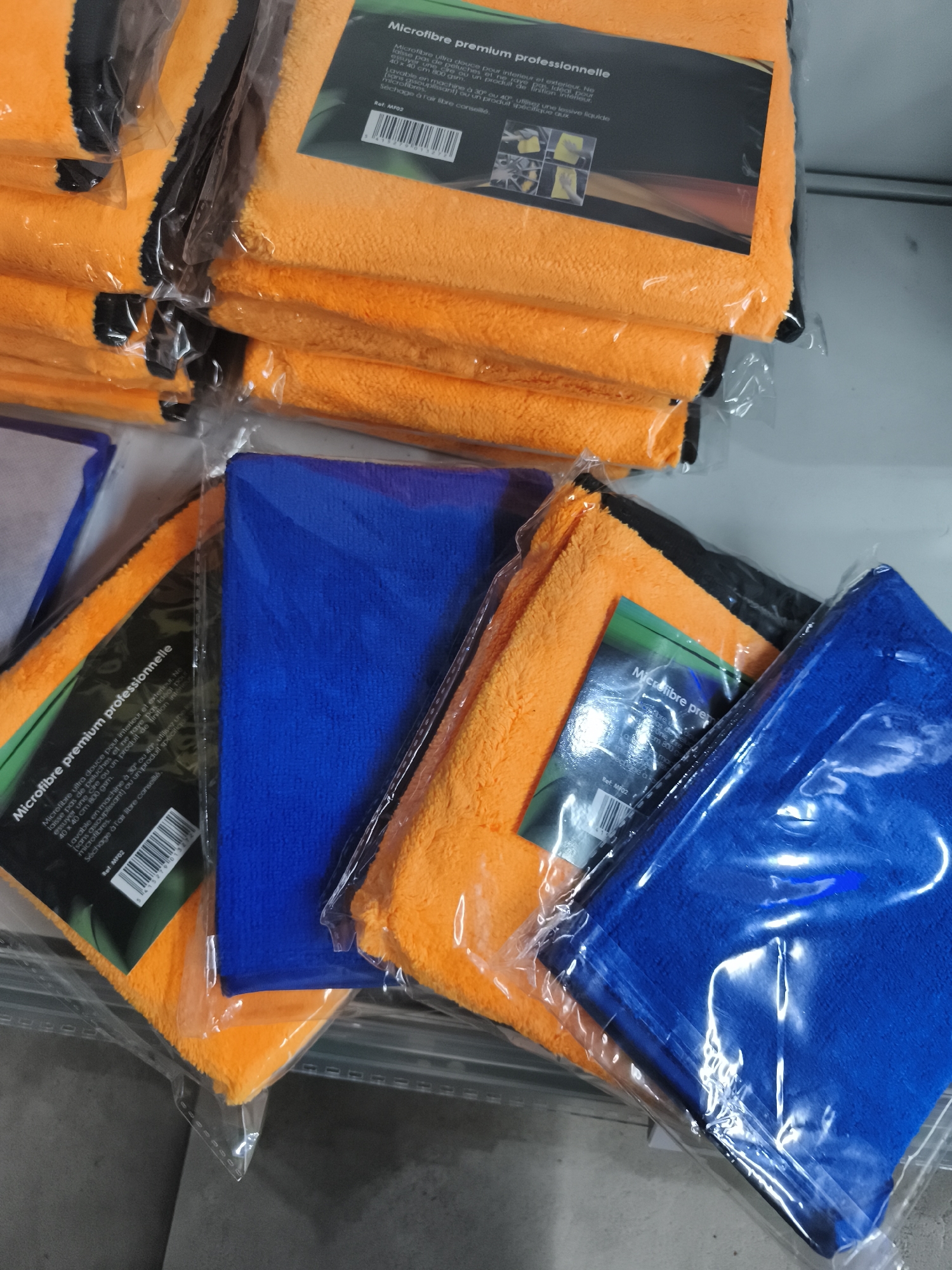 The image shows a retail shelf in Saint-Denis, France, displaying stacks of packaged microfiber cloths. The cloths are predominantly orange and blue. A label on one of the orange cloth packages reads "Microfibre premium professionnelle" and includes care instructions and a barcode. The cloths are neatly folded and appear to be new, sealed in clear plastic packaging. The shelf itself is metal and appears to be in a store or warehouse setting.