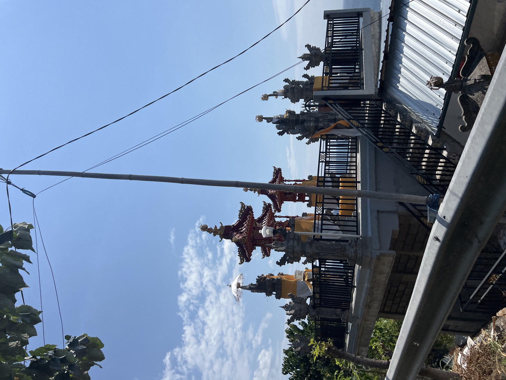 The image depicts an outdoor scene in Abang, Indonesia, featuring an elevated platform adorned with Balinese temple architecture. Several multi-tiered red and gold structures, characteristic of Balinese shrines, are visible on the platform, some topped with golden finials. Stone carvings, some with yellow and white fabric draped around them, punctuate the railing of the platform. A white ceremonial umbrella is prominently displayed on one of the stone structures. A black metal fence borders the platform, and a set of black metal stairs leads down from it. The background reveals a bright blue sky with scattered white clouds and a hint of the ocean in the distance. To the left, lush green trees and foliage frame the scene, and to the right, a corrugated metal roof is visible. In the foreground, a grey metal barrier runs horizontally across the image. A utility pole with electrical wires stretches vertically through the composition, partially obscuring the sky and temple structures. No people are visible in the image.
