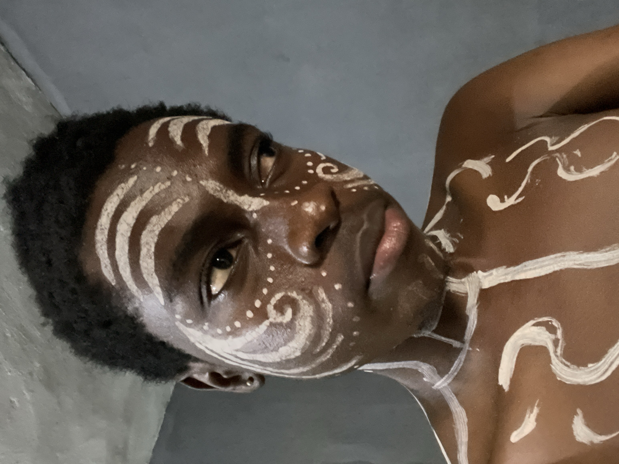 A close-up, rotated portrait features a person with dark skin, adorned with intricate white body paint on their face and upper body. The subject is positioned diagonally in the frame, looking upwards and slightly to the right with a neutral and contemplative expression. The image, taken in Nkpor, Nigeria, captures the subject against a plain, dark grey background, indicative of an indoor setting.
The white body paint forms distinct patterns across the forehead, temples, cheekbones, and nose. These patterns include bold, curved stripes on the forehead, small dots around the eyes and on the nose, and spiraling motifs on the cheek and temple. Similar white lines and swirling designs extend onto the visible shoulder and chest area, indicating extensive body art.
The subject has short, dark, curly hair that frames their face. Their lips are visible, appearing full. The lighting highlights the texture of the skin and the raised application of the white paint. No text is visible in the image. The absence of outdoor elements or specific time-of-day cues suggests a controlled environment, possibly for a cultural performance, artistic expression, or photoshoot.