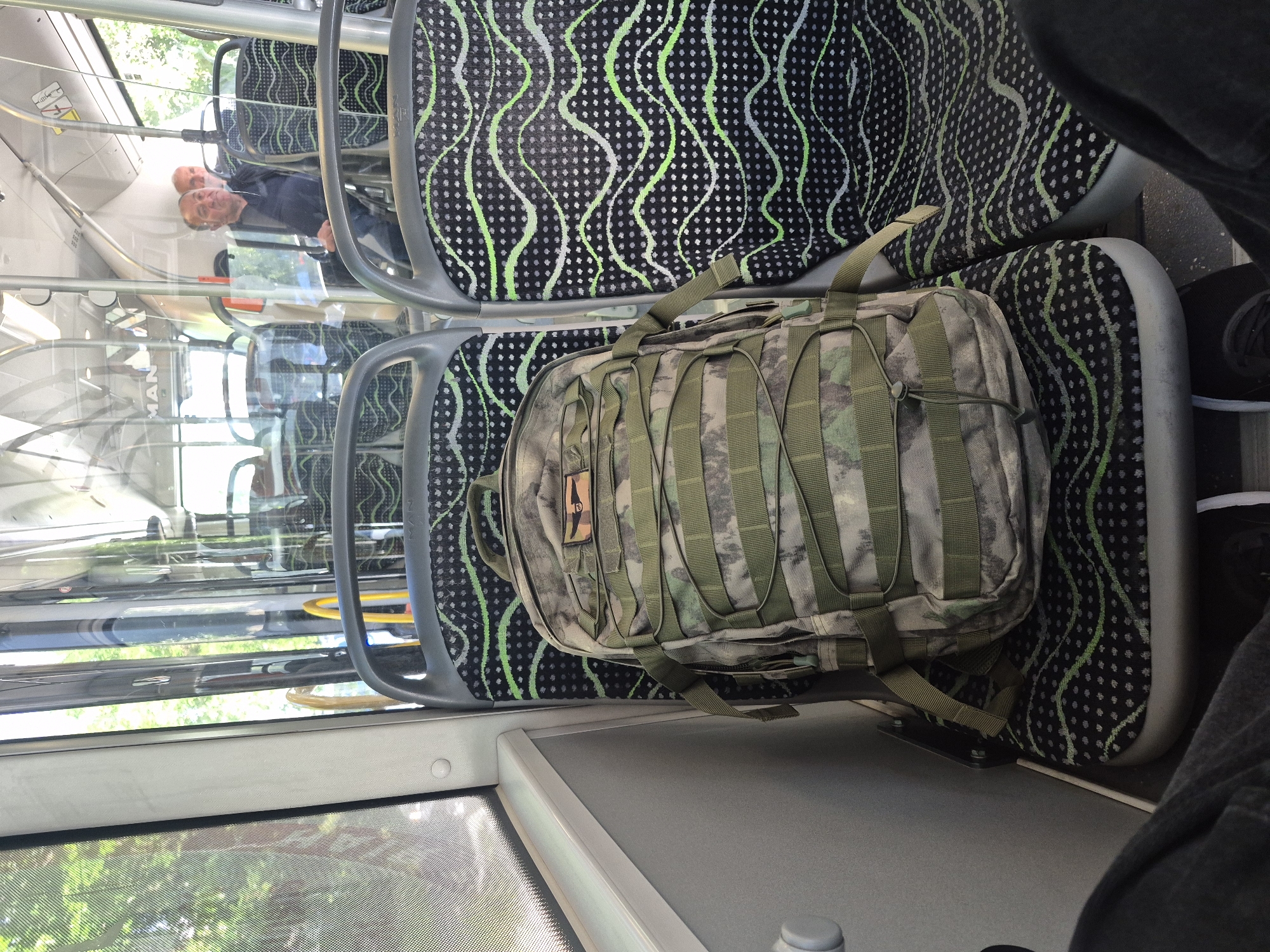 The image shows the interior of a bus, likely in Tbilisi, Georgia. A camouflage backpack is resting on a seat with black fabric featuring wavy green and white lines over a pattern of gray dots. Visible through the bus windows are trees and sunlight, indicating an outdoor environment. A man's reflection can be seen in the window, wearing a dark jacket. Another person is partially visible in the lower right, wearing a gray garment and black and white shoes.