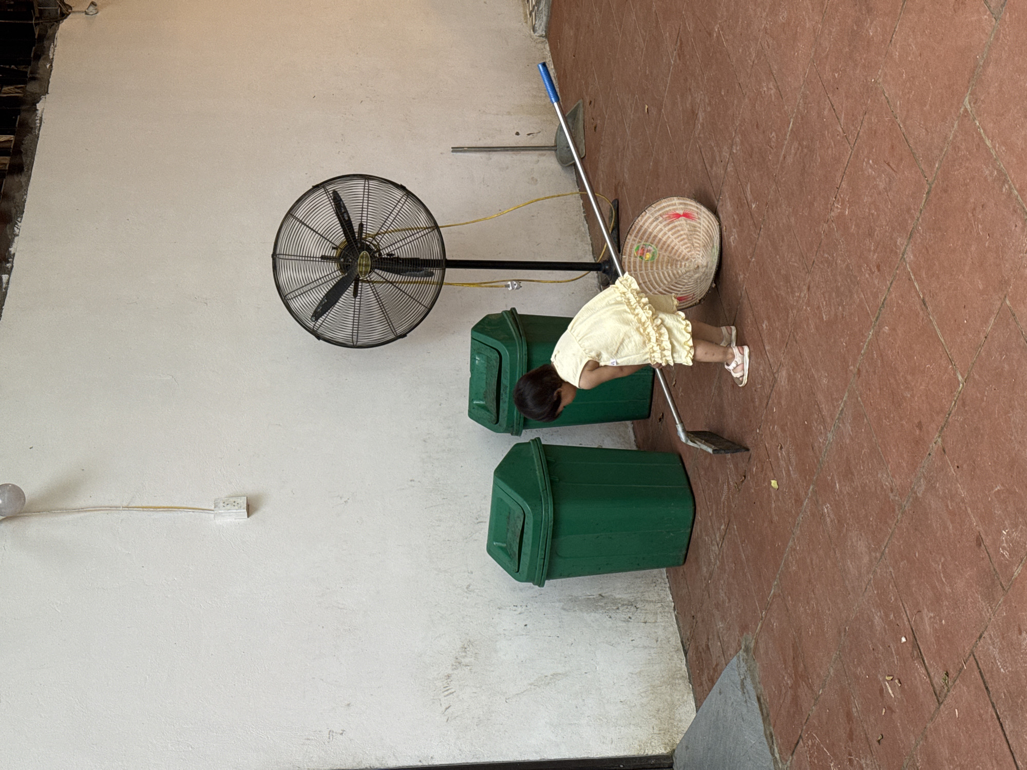 A young child in a yellow dress and sandals is sweeping a tiled floor with a broom. A traditional conical hat lies on its side on the floor near the child. In the background, two green trash cans stand against a white wall. A large industrial fan on a stand is also visible, along with a dustpan and broom leaning against the wall. The scene appears to be an outdoor covered area, possibly a patio or walkway. The location is identified as Cổ Lũng Commune, Vietnam.