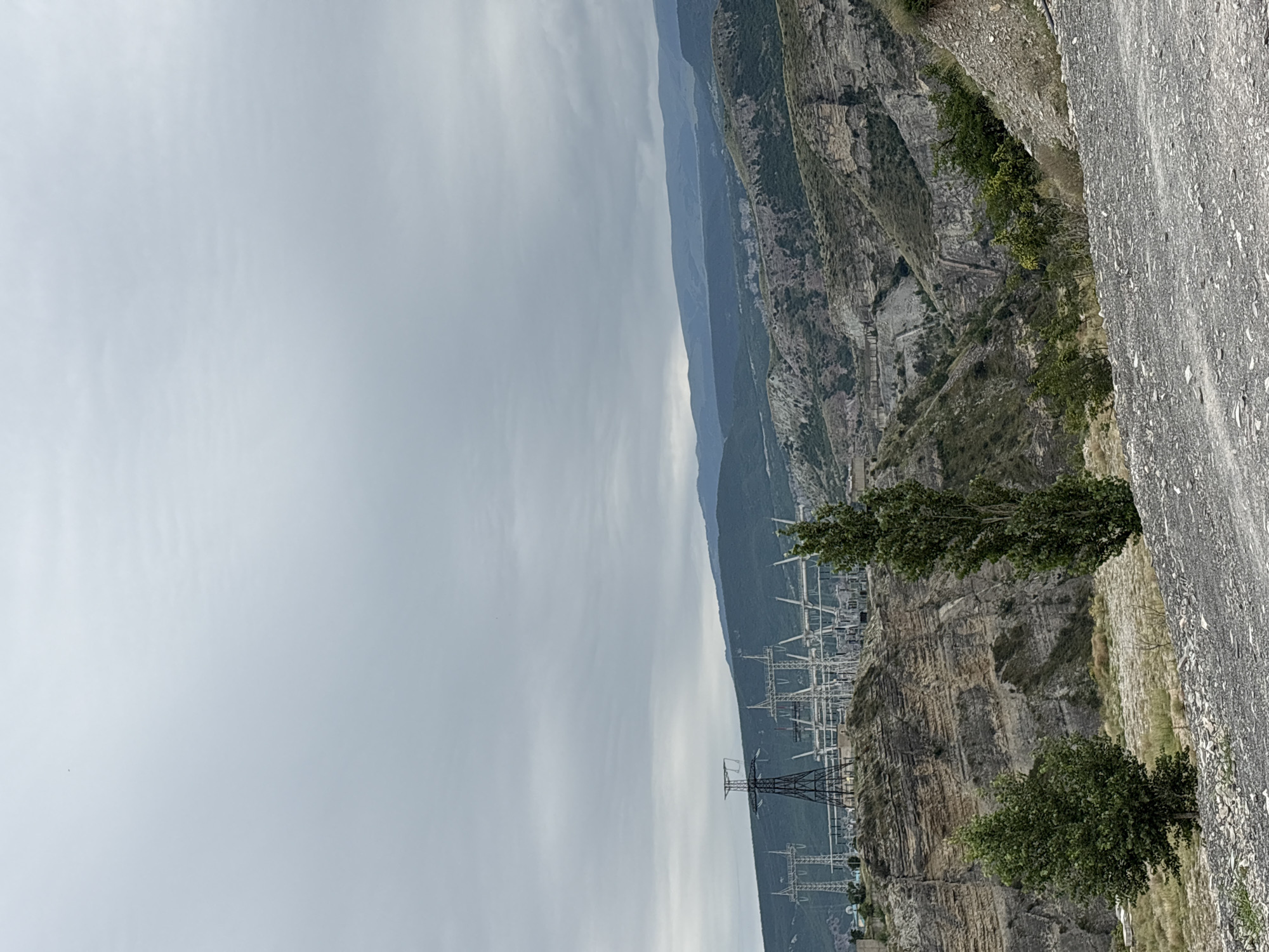 This is a photograph of the Chirkey Hydroelectric Power Station in Chirkey, Russia. The power station is situated in a mountainous, rugged landscape. In the foreground, a gravel road leads the viewer's eye towards a vast, layered rock formation on the left side of the image. Two trees, one smaller and bushier on the left and a taller, slender one on the right, stand in front of the rock face.
In the midground, the imposing structures of the Chirkey Hydroelectric Power Station are visible. Large power lines supported by tall metal towers stretch across the scene. The station appears to be built into the side of the mountain. The background reveals a series of rolling hills and mountains, covered in vegetation and shrouded in a hazy, overcast sky. The overall impression is one of industrial infrastructure integrated into a dramatic natural setting.