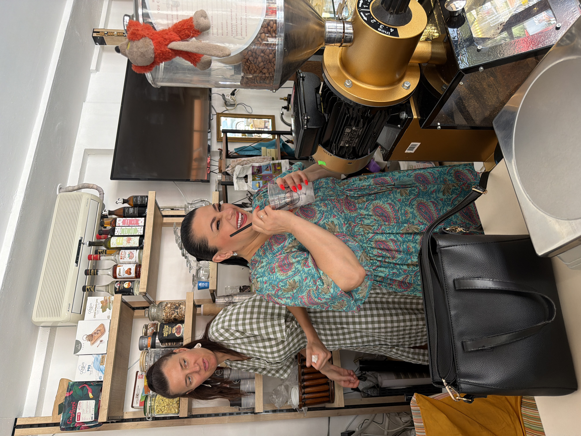 In Marmaris, Turkey, two women are visible inside a cafe, standing behind a counter. The woman on the right, wearing a patterned dress, is laughing and holding a drink with a straw. She has dark hair and is wearing earrings and a ring. The woman on the left, wearing a green checkered dress, is looking away from the camera with an exaggerated expression. She has her hair pulled back and is wearing earrings. In the background, shelves are stocked with various products, including jars of food, boxes of tea, and bottles of syrup. A large coffee grinder, a television screen, and a stuffed fox toy are also present. A black handbag is placed on the counter in the foreground.