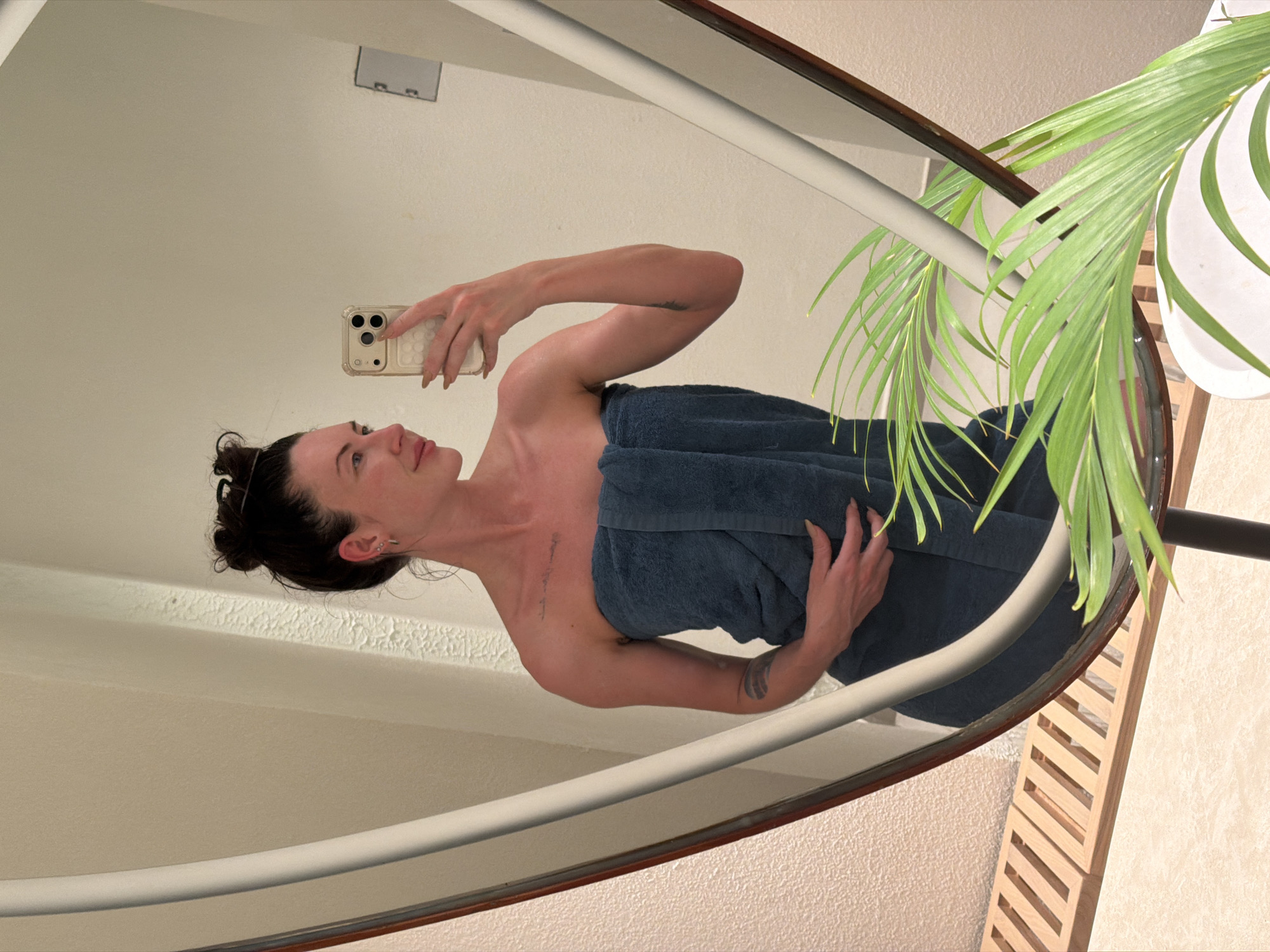 A woman is taking a mirror selfie. She is wearing a dark blue towel wrapped around her body. She has her hair up in a bun. Her left arm is raised, holding a phone. She is wearing earrings and has a tattoo on her left arm and chest. The background appears to be a bathroom with a textured wall, a wooden slatted shelf, and a plant in the foreground. The image does not provide any specific details about the city of Dong Ninh Hoa Ward, Vietnam.