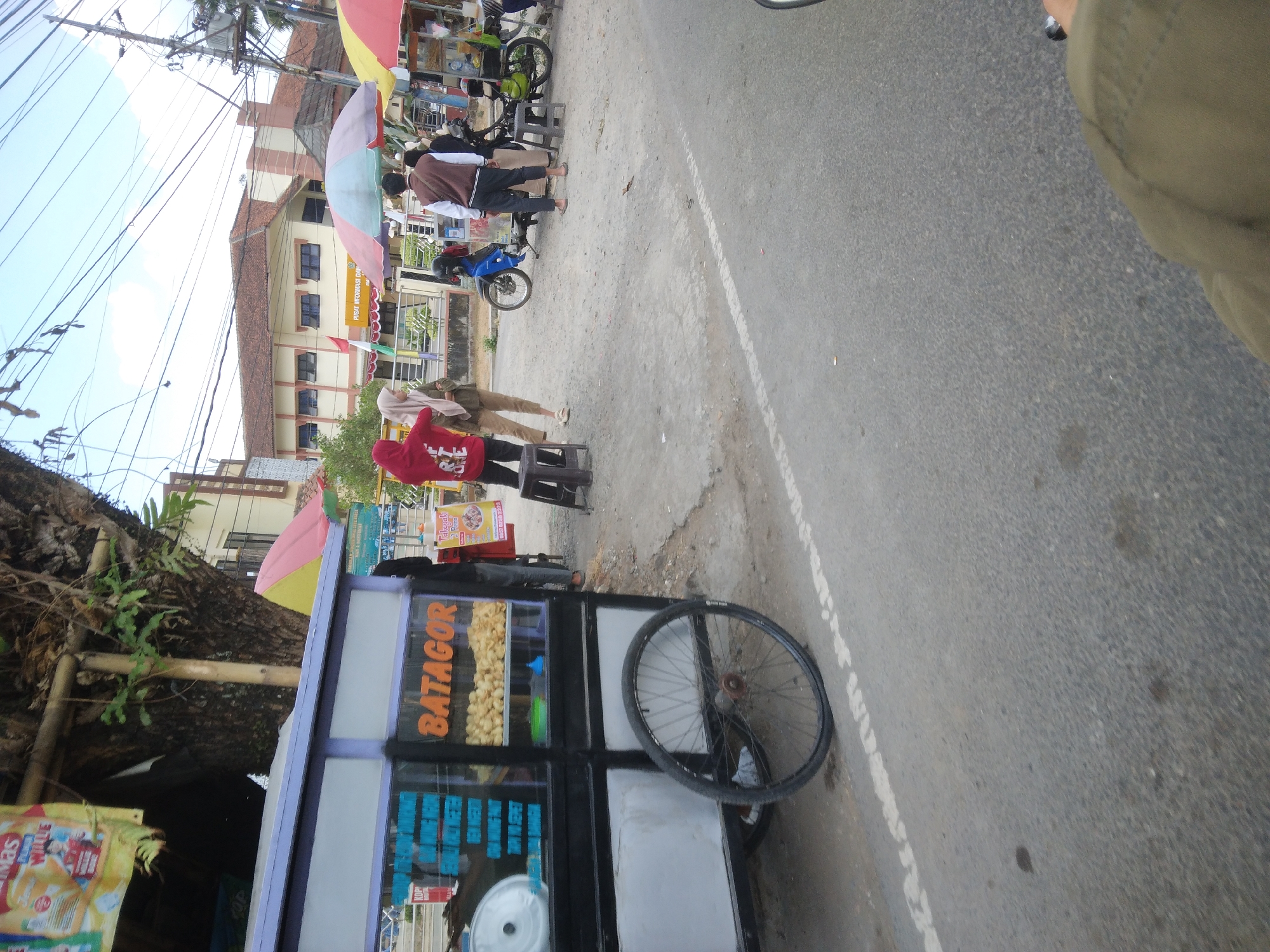 The image depicts a street scene in Berkoh, Indonesia. In the foreground, a mobile food cart with the word "BATAGOR" prominently displayed is parked on the side of a paved road. The cart has a blue and white exterior and contains displayed food items, likely Batagor, a type of fried snack. Next to the cart, a large spoked wheel from the cart is visible. Several individuals are present near the food cart and in the background. Two people are standing by the cart, one in a red hoodie and the other in a beige hijab and outfit. Further back, two men are standing near motorcycles, one in a white and brown jacket and the other in a black jacket. In the background, a building with a tiled roof and several windows is visible. The scene is outdoors with overhead power lines and a cloudy sky. A portion of the image also shows the leg of a person wearing a green jacket, suggesting the photo was taken from a seated perspective, possibly on a motorcycle.
