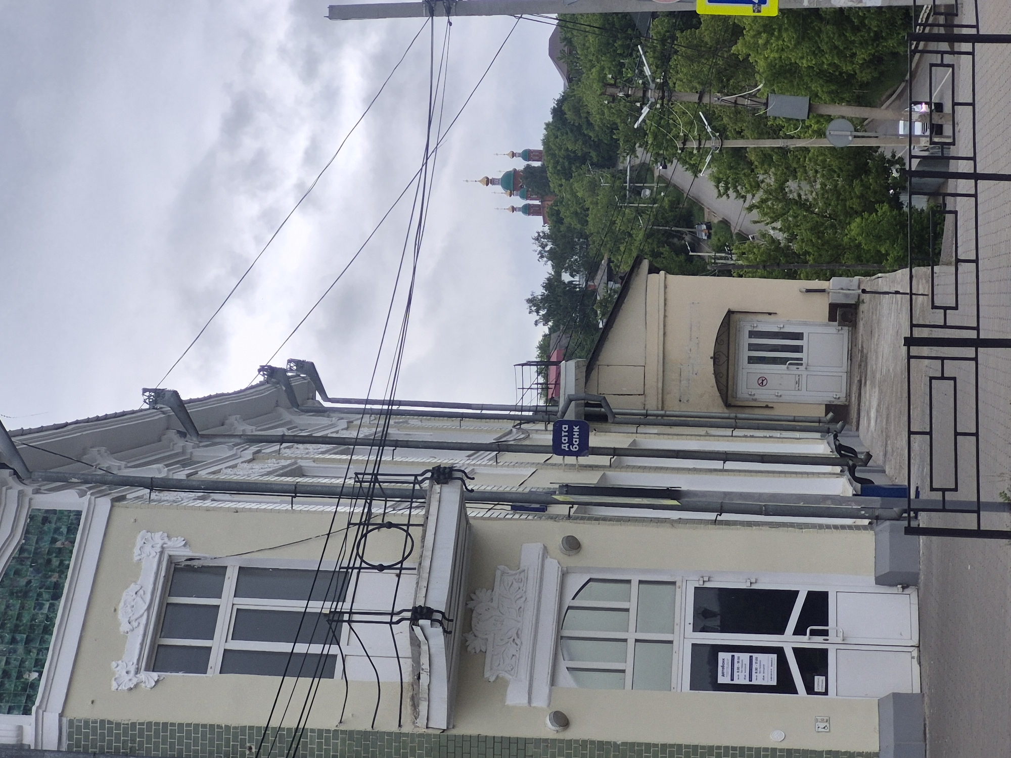 This image depicts a street scene in Sarapul, Udmurt Republic, Russia, specifically looking southeast from the vicinity of Krasnoarmeyskaya Ulitsa, 70.
The left side of the frame is dominated by a light yellow, multi-story building identified as "Дата Банк" (Data Bank) by a prominent blue sign with white Cyrillic text. This building features elaborate white architectural ornamentation around its windows and a small balcony with a black wrought-iron railing. The upper left facade includes a section of green square tiles. A double glass door with white frames, displaying informational signs and an "X"-shaped tape, serves as the main entrance on the ground floor. Further to the right along the building's facade, another white door with vertical panels displays a "no smoking" symbol. Several black utility wires stretch across the upper and mid-sections of the image, originating from poles partially visible on the right.
To the right of the building, a paved road, likely Nagornaya Ulitsa, descends into the distance, bordered by dense green deciduous trees. Multiple utility poles equipped with streetlights line this winding road. Two vehicles are visible: a light-colored car with illuminated headlights in the lower right, and another vehicle further up the road. A yellow pedestrian crossing sign is partially visible on the far right.
In the background, atop a tree-covered hill, stands the Church of the Beheading of John the Baptist (Церковь Усекновения Главы Иоанна Предтечи). This church is characterized by its red brick structure and multiple vibrant green domes, each topped with a golden cupola and an Orthodox cross. The sky above is uniformly overcast with light gray clouds. In the immediate foreground, a black metal fence with a pattern of connected rectangles separates the viewer from the downward sloping street.