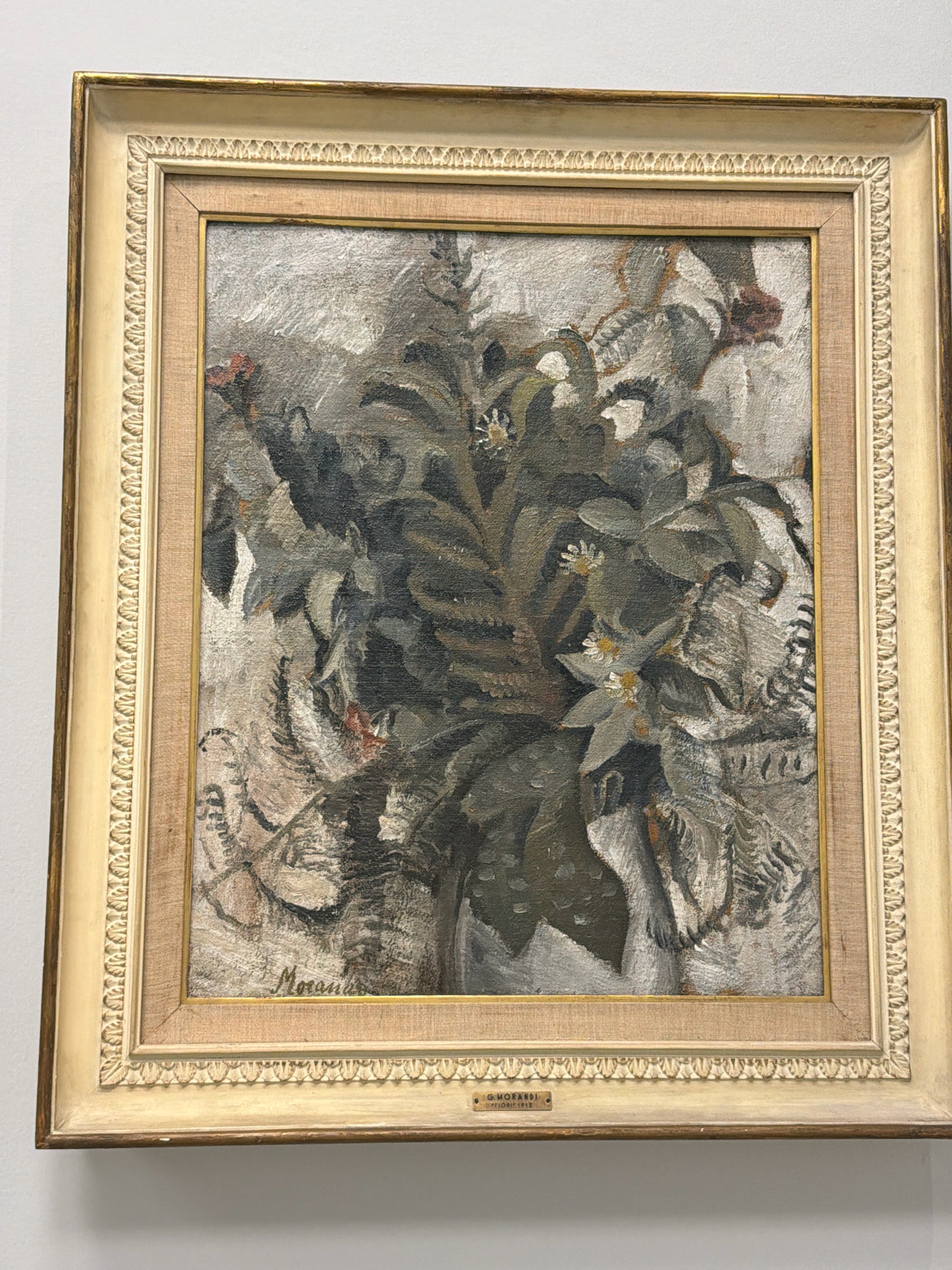 The image shows a framed painting hanging on a white wall. The painting itself is an oil on canvas still life of flowers and foliage in muted tones of gray, green, and white, with some hints of red. The brushwork appears to be thick and textured. The artist's signature, "Morandi," is visible in the lower left of the painting. A small plaque beneath the painting reads "G. MORANDI / SETTIMO 1913". This painting is located in the Galleria d'Arte Moderna (Gallery of Modern Art) in Milan, Italy.