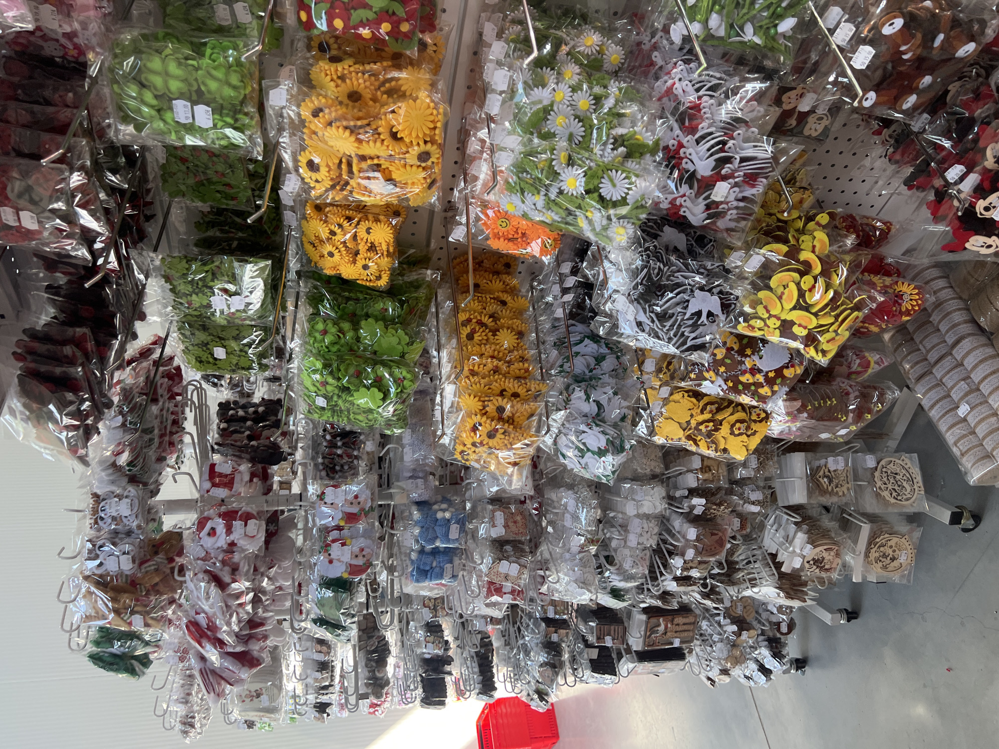 The image depicts a rack of small, packaged crafting supplies for sale in a retail setting. The packs include items such as flower decorations, shapes, and other decorative components, suspended from metal hooks on a metal rack. The location is presumed to be a retail store or craft shop possibly located in Burgas, Bulgaria. There are no people depicted. The environment suggests an indoor retail setting with a tiled floor.