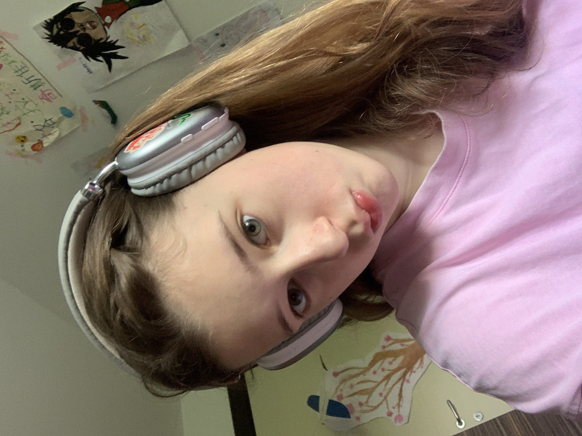 A young person with light skin and long, light brown hair is wearing headphones and a pink shirt. They are looking directly at the camera with a slight pout. The background is a light-colored wall with drawings and papers attached. A picture of a cartoon character is visible. The location is indoors. No location in Vozdovac Urban Municipality, Serbia is provided or confirmed.