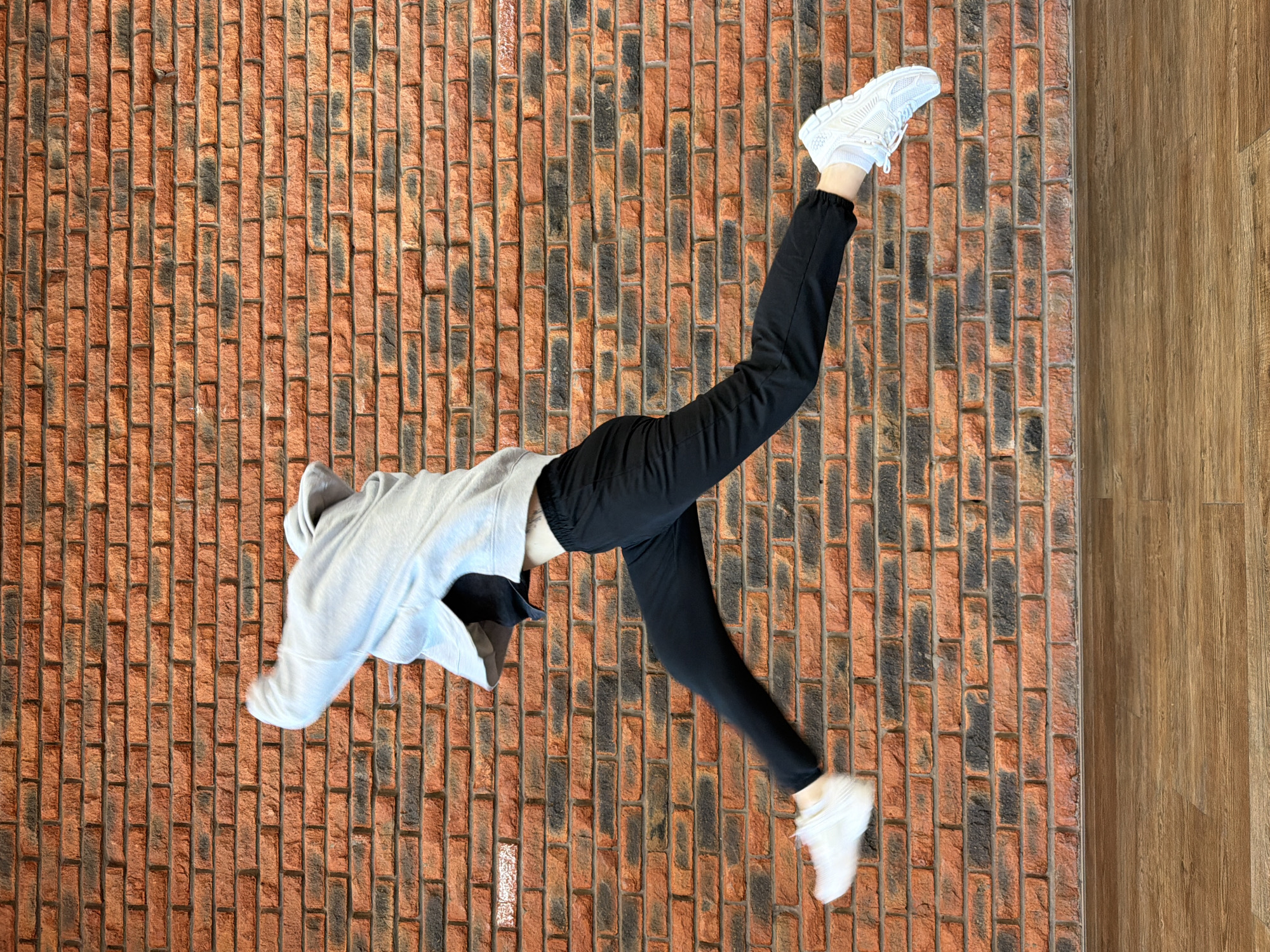 A person in a grey hoodie, black pants, and white shoes is positioned in a horizontal mid-air split, lying on a floor that transitions from a red brick pattern on the left to a wooden pattern on the right. The setting appears to be indoors, possibly a studio or a room with a distinct flooring design. There is no clear information about the city of Chaikovsky, Russia.