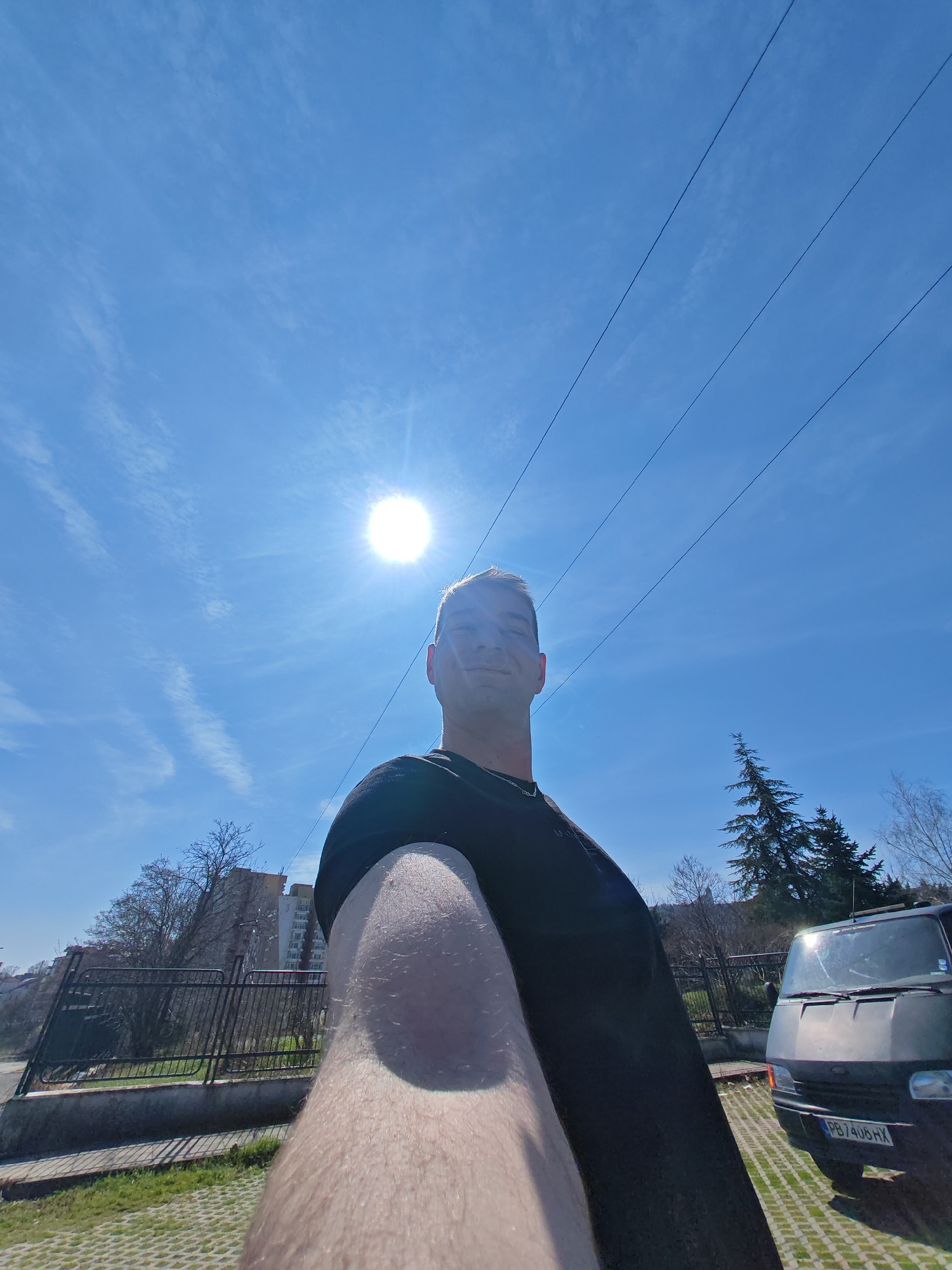 The image depicts an outdoor daytime scene in the city of Shumen, Bulgaria, under a clear blue sky with sparse, wispy clouds and a prominent sunburst in the upper left quadrant. Three parallel overhead power lines traverse the upper portion of the sky.
A male subject is prominently featured in the lower-center, appearing to take a selfie. His head and extended right arm (from the shoulder to the elbow) are visible, filling a significant portion of the lower frame. He has short, light-colored hair and is wearing a dark t-shirt. A faint smile is discernible.
In the mid-ground, behind the subject's arm, a black metal fence with vertical bars and a horizontal top rail runs across the scene. Beyond the fence, a sloped area features mostly bare deciduous trees and a tall, dark green coniferous tree. Multi-story residential buildings, one light-colored, are visible in the background on the left, nestled among the trees.
To the right of the subject, a dark-colored (likely black or dark grey) Ford Transit van is parked on a permeable paver surface. The front of the van is visible, displaying a Bulgarian license plate: "PB 7406 HX." The ground in the foreground and beneath the van is composed of grid-patterned pavers with green grass growing through the openings, indicating a parking area. The scene is brightly lit by direct sunlight, casting some shadows.