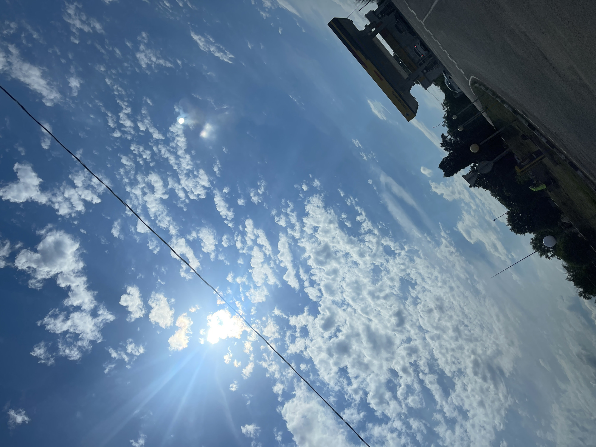 The image depicts a bright, sunny sky with scattered white cumulus clouds, suggesting fair weather. The sun is visible, casting strong rays and creating a lens flare effect, indicating it is likely daytime, possibly mid-morning or early afternoon. A black wire stretches diagonally across the sky, suggesting the presence of overhead electrical lines or communication cables.
In the upper right quadrant of the image, a portion of a road or paved area is visible, along with some trees and lampposts, hinting at an outdoor, possibly urban or suburban, setting. A yellow structure, possibly part of a building or signage, is also present. A car is visible in the distance.
The overall scene is dominated by the vast expanse of the sky, conveying a sense of openness and natural beauty. There is no clear activity or event taking place, and no people are visible. The rotation of the image suggests it was taken with the camera held at an angle.
No discernible text is visible in the image. The location context of Donskoe, Russia, places this scene within that geographic area.