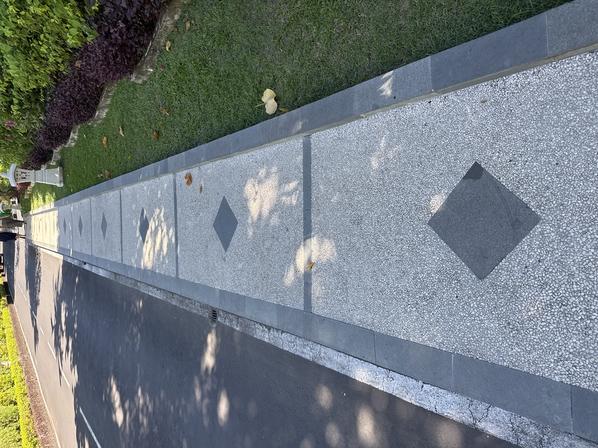 This elevated view captures a well-maintained outdoor scene in Denpasar, Indonesia, focusing on a pedestrian walkway, a road, and surrounding greenery under clear, sunny conditions.
The central subject is a long, light-colored pathway, appearing to be made of pebble-dash or small aggregate, with a textured, speckled surface. This walkway is intermittently broken by dark grey square inserts, rotated to form a diamond pattern, creating a repetitive geometric design along its length. A dark grey curb neatly separates the walkway from the adjacent areas.
To the left (top of the image), a strip of vibrant green grass is visible, with a few yellowish-brown fallen leaves scattered upon it. Beyond the grass, there is a dense border of lush, dark purple-leafed plants, which then gives way to brighter green foliage further in the background. A decorative white stone lantern or similar garden ornament is partially obscured by the plants near the start of this section.
To the right (bottom of the image), an asphalt road runs parallel to the walkway. The edge of the road features a white painted line, indicating a boundary or lane marking. Numerous dappled shadows of tree leaves are cast across both the walkway and the road, suggesting bright sunlight from an angled source, typical of morning or late afternoon.
No people are visible in the image, and no specific activities are taking place, creating a quiet and orderly impression of a public or park area. There is no visible text.