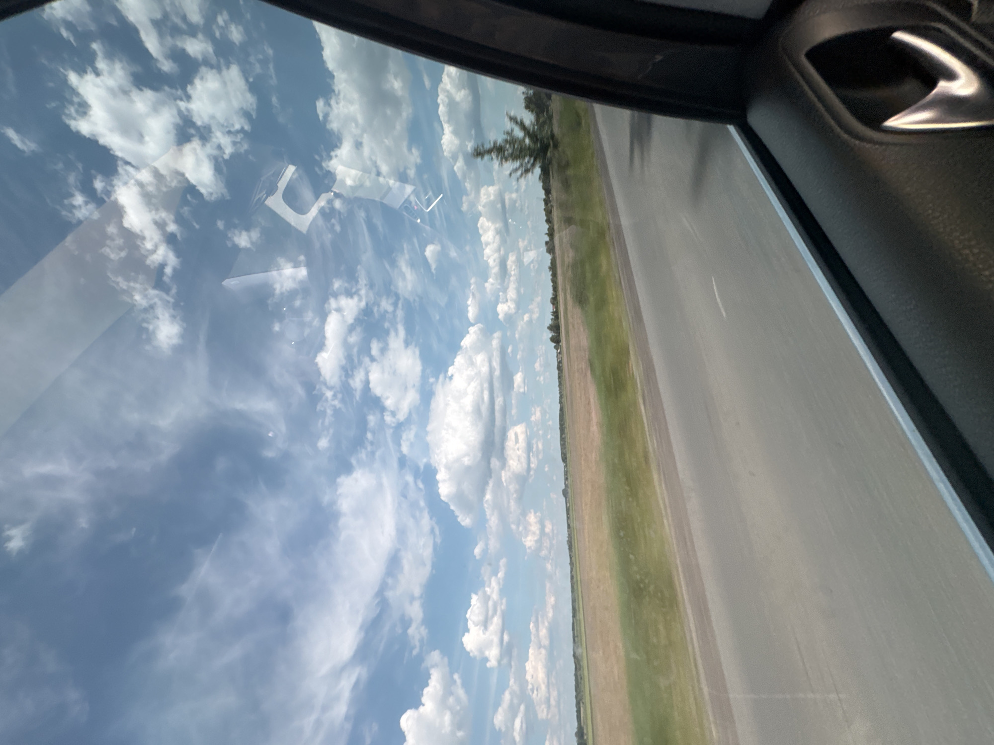 The image displays a rural landscape near Markivka, Ukraine, observed from the interior of a moving vehicle, specifically through a passenger-side window. The upper two-thirds of the scene feature a bright blue sky populated by numerous white cumulus clouds and scattered wispy cirrus clouds. Reflections of the sky and interior elements are visible on the window glass, particularly in the upper right quadrant.
Below the sky, a two-lane asphalt road extends horizontally across the middle to lower sections of the image, showing faint white dashed lines in the distance and suggesting forward motion due to slight blur in the foreground. Adjacent to the road, extensive flat agricultural fields are visible; some sections appear as tilled, brownish soil, while others are covered with green vegetation. A distant treeline defines the horizon, with barely discernible structures visible beyond it. A single evergreen tree, possibly a pine or fir, stands closer to the roadside on the right side of the landscape, exhibiting subtle motion blur.
The lower right portion of the image reveals a segment of the vehicle's dark gray, textured interior door panel, including a reflective, metallic door handle and a narrow silver-colored window trim. No individuals or animals are present in the captured environment. The scene is illuminated by bright daylight.