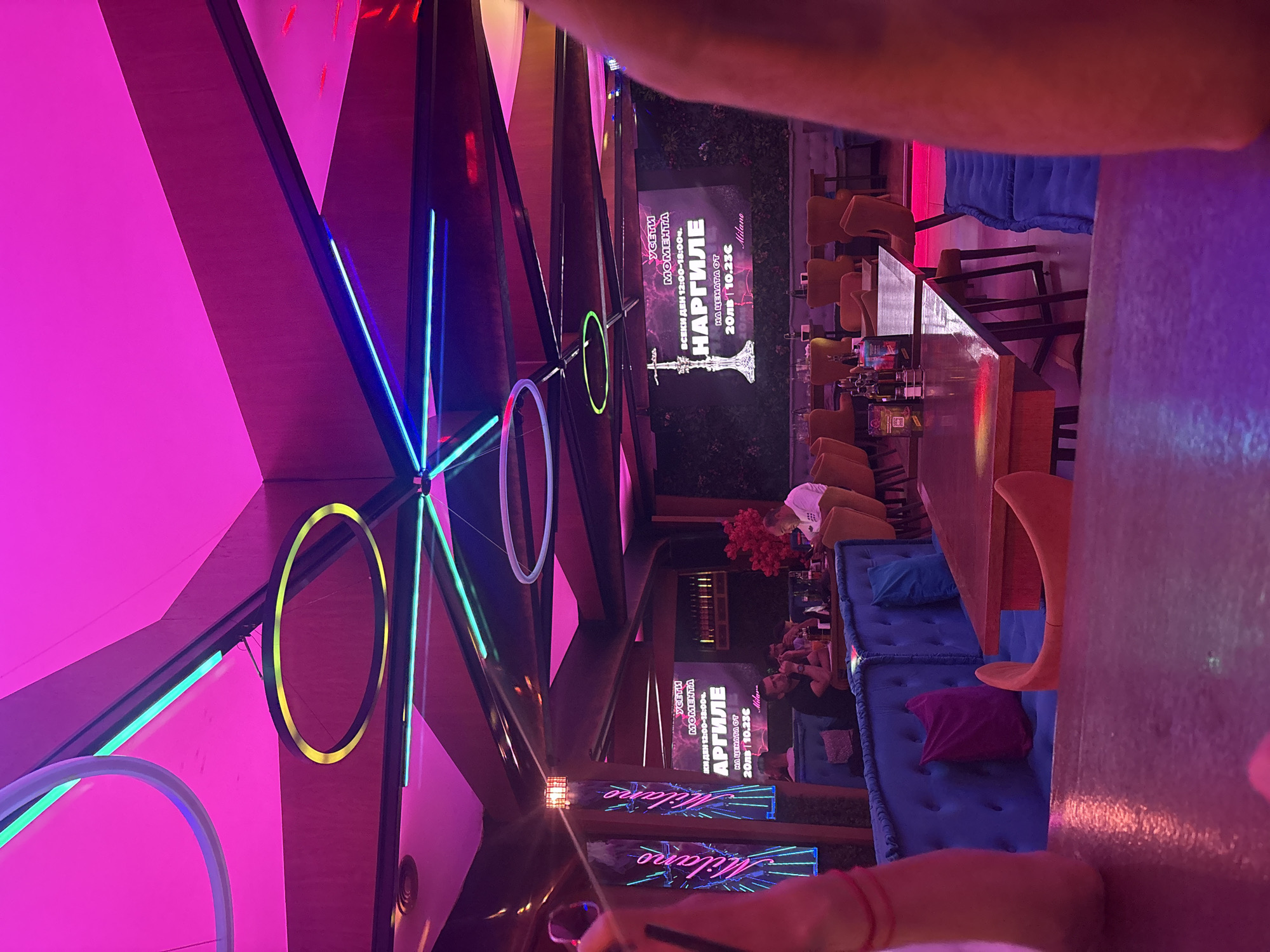The image depicts the interior of a dimly lit, modern establishment, possibly a lounge or bar, with a prominent pink and purple neon lighting scheme. Geometric patterns are visible on the ceiling, illuminated by colorful LED strips and circular fixtures. Several tables are set up for seating, with blue upholstered benches and individual orange chairs. In the background, patrons are seated at tables, engaged in conversations or observing their surroundings. A large screen displays an advertisement featuring a hookah, with text in what appears to be Bulgarian, indicating "Усети момента" (Feel the moment) and details about "Наргиле" (Hookah) pricing. Two vertical neon signs with the word "Milano" are also visible on the left side of the image. The overall ambiance suggests a contemporary nightlife venue.