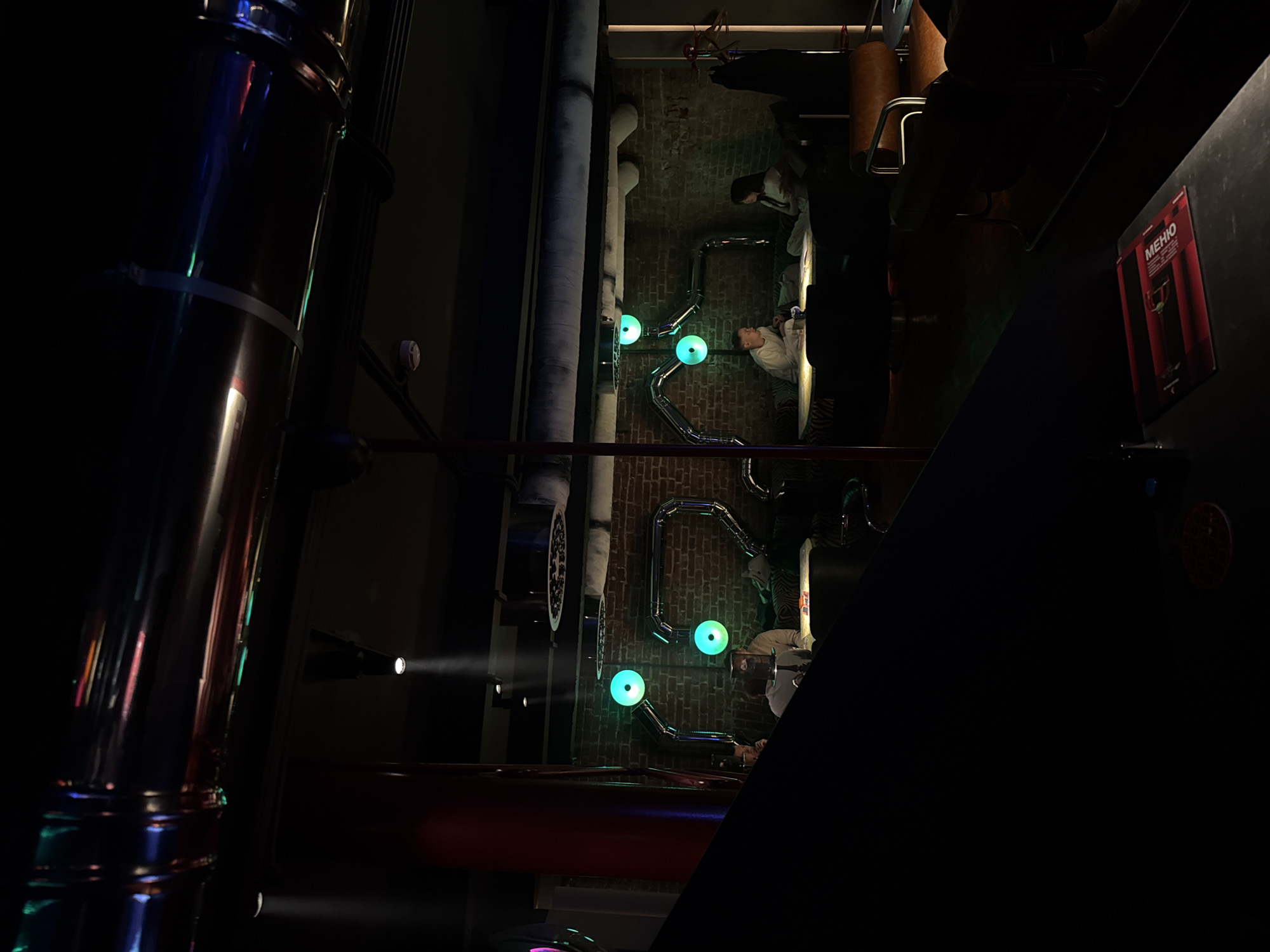 The image shows the interior of a restaurant in Moscow, Russia. The scene is dimly lit, with artificial light sources highlighting specific areas. Several people are seated at tables, seemingly engaged in conversation or dining. The décor includes exposed pipes with decorative green lights. A sign with the text "MEHIO" is visible on a nearby wall. A shiny metallic column is prominent in the foreground. The overall atmosphere appears to be modern and industrial-chic.