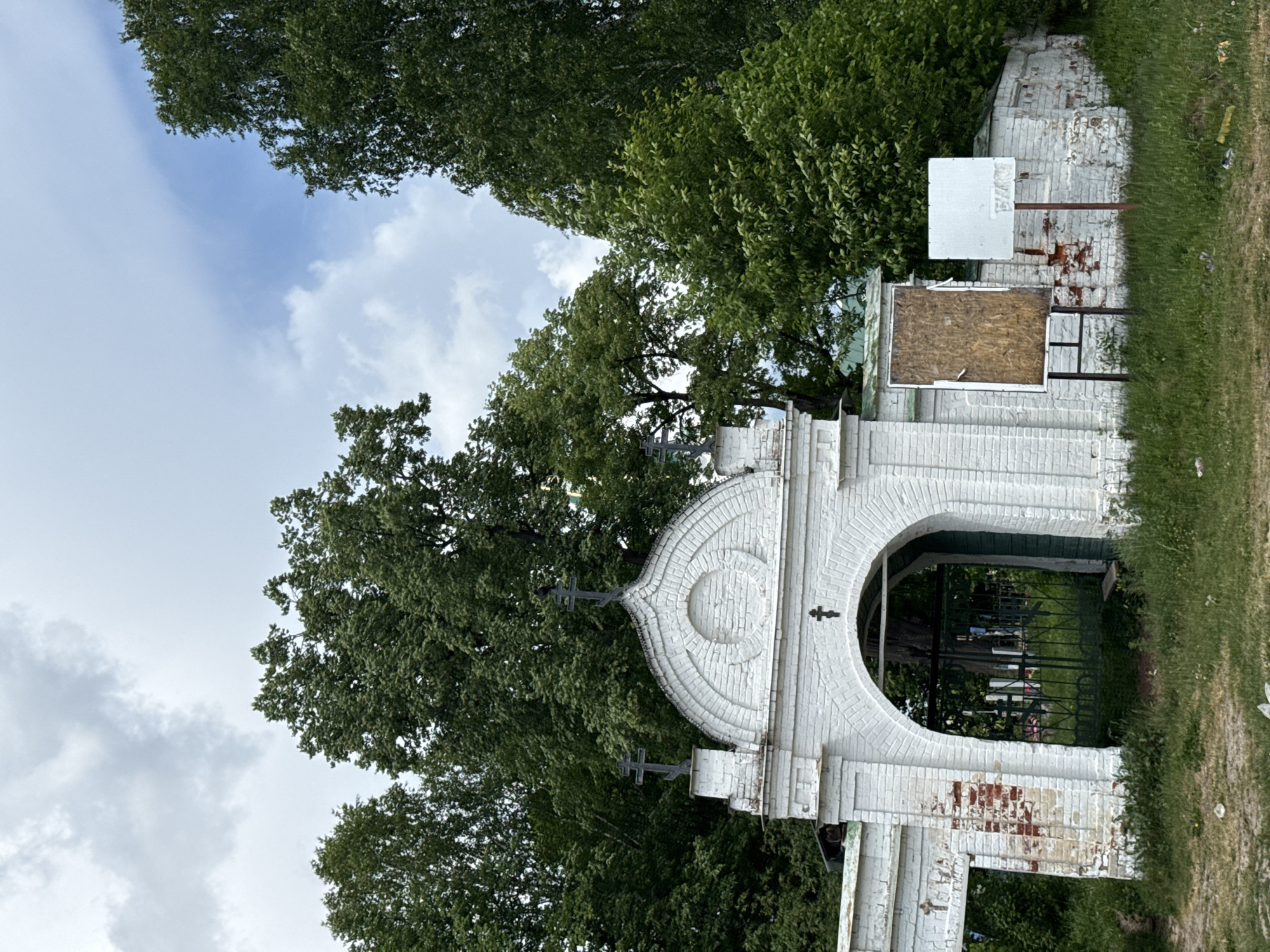 The image shows a white brick archway with crosses on top, possibly the entrance to a cemetery, surrounded by trees and greenery. The archway appears aged, with some brick exposed. In the background, through the archway, is a glimpse of the cemetery, with some grave markers visible. There are signs on the right side of the archway, one a blank white rectangle, the other a blank rectangle of wood. The sky above is cloudy. The location is assumed to be in or near the city of Chistoe Pole (Lindovskii S/S), Russia.