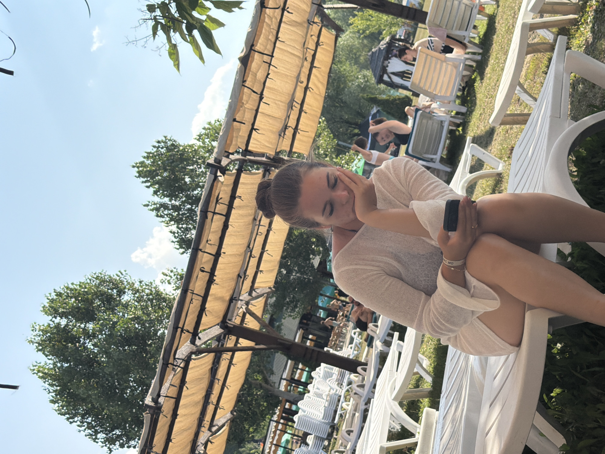 A young Caucasian woman with her hair in a bun sits on a white lounge chair at an outdoor resort. She is wearing a sheer white cover-up over a black swimsuit and is holding a black smartphone in her right hand, with her chin resting on her left hand. She is looking down at the phone with her eyes closed. In the background, there are rows of empty white lounge chairs, trees, and people relaxing. The scene appears to be set during the daytime, under a clear blue sky with scattered clouds. The location is identified as Sociteni, Moldova.