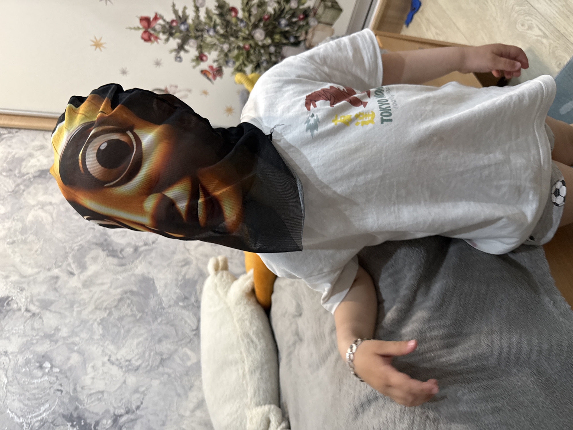 A child wearing a mask is lying on a gray cushion, with another white cushion next to it. The child is wearing a white t-shirt with "TOKYO" printed on it, and gray shorts with a soccer ball design. The environment includes a light-colored floor and wall, with a plant visible in the background. The location is Rusanovo, Russia.