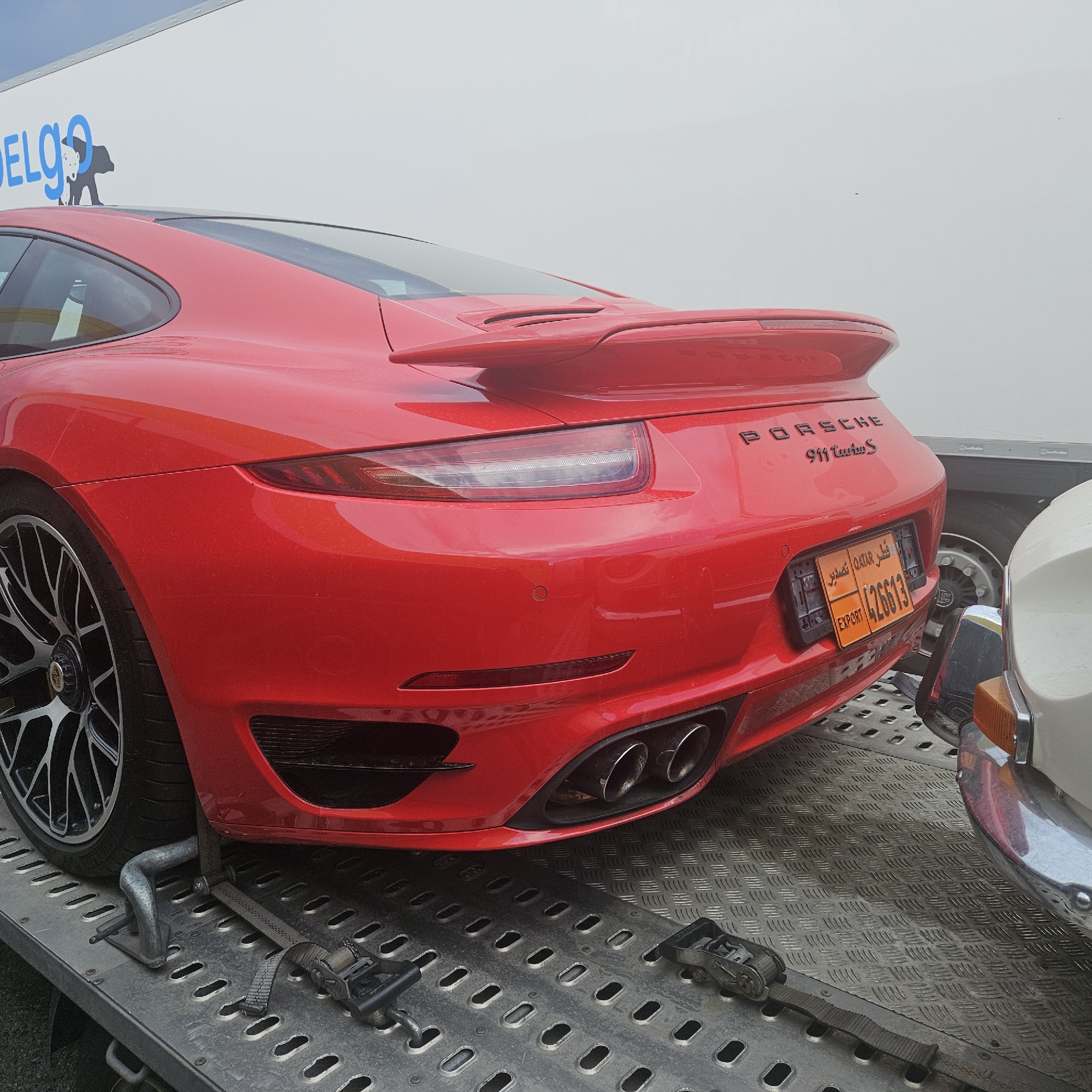 In La Jonquera, Spain, a red Porsche 911 Turbo S is secured on a trailer. The car's rear is visible, featuring its taillights, quad exhaust pipes, and a black spoiler. The license plate is orange with Arabic and English text and the number "426613". Straps and metal securing mechanisms are in place to prevent the car from moving. The trailer is made of metal with a diamond plate pattern. In the background, a white truck with a logo partially visible, reading "DELGO" and a silhouette of a bear, is parked. Another vehicle, partially visible on the right, appears to be a classic car with a chrome bumper and an amber turn signal. The environment suggests an outdoor loading or transport area.