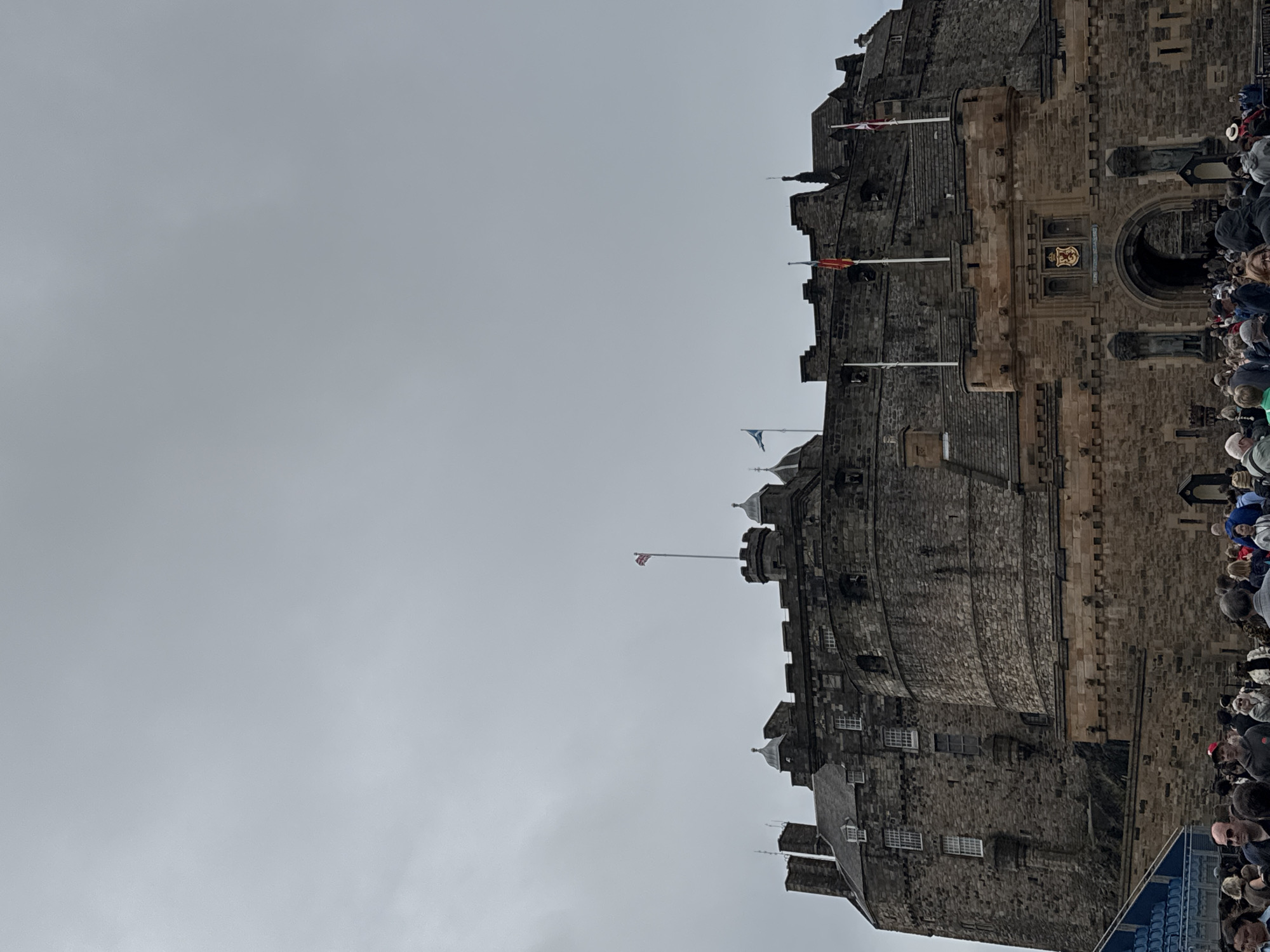 The image captures a low-angle view of Edinburgh Castle, a historic fortress perched on Castle Rock in the city of Edinburgh, Scotland, United Kingdom. The castle, constructed from rough-hewn stone, dominates the upper portion of the frame. Its battlements, turrets, and arched gateways are visible, with flags flying from several points on the structure. The sky above is overcast and a pale grey. At the base of the castle, a crowd of tourists is gathered, looking towards the entrance. The people are diverse in age and attire, some holding cameras, suggesting they are observing or entering the castle. In the lower left corner, bleachers or seating can be seen, partially obscured by people. The overall scene depicts a popular tourist attraction on a cloudy day, with a significant number of visitors.