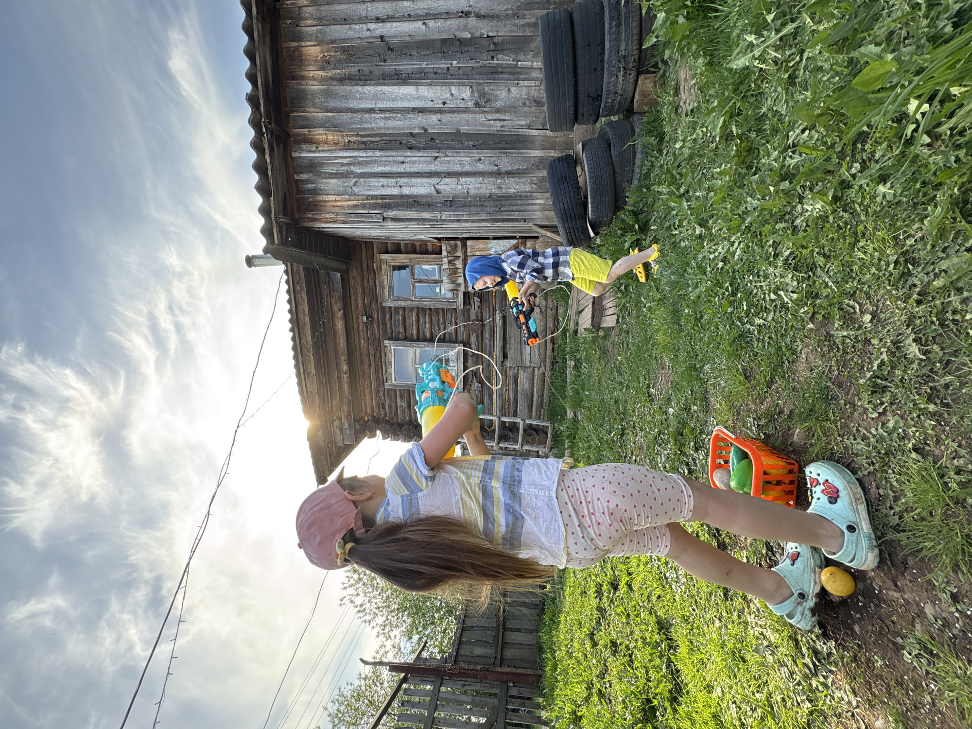 The image depicts two children engaged in play in a rural environment, likely in the vicinity of Nizhnyaya Chermoda, Russia, as specified. The older child, a girl with long brown hair wearing a pink cap, a striped shirt, and polka-dot shorts, stands with a water gun in her hands. Near her feet is an orange basket containing various objects, possibly toys or vegetables, and she wears blue sandals. Nearby lies a yellow object, possibly a potato or lemon.
In the background, a younger child, a boy wearing a checkered shirt and yellow shorts, also holds a water gun. He is positioned near a weathered wooden building with stacked tires leaning against its exterior wall. The environment is grassy and unkempt, with evidence of weeds and uncultivated vegetation. The sky overhead is partially cloudy, indicating daylight.