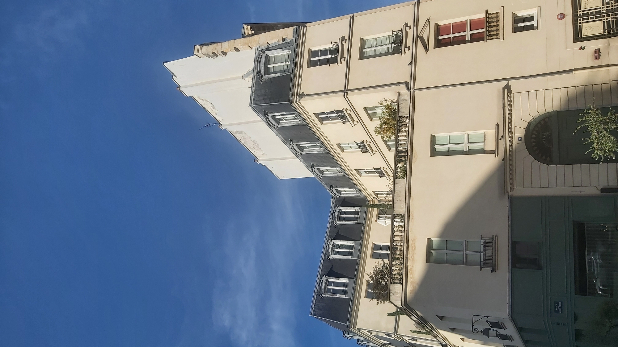 This is a low-angle, full-shot photograph of a building with a mansard roof against a clear blue sky with wispy clouds. The building is pale yellow with dark grey mansard roofs, dotted with dormer windows. The structure appears to be an apartment building or hotel, with several balconies and windows visible. The lower part of the building on the left has a green facade with some signage, including a street name sign reading "PLACE PIERRE FUMEY". On the right side of the building, there is a large arched doorway with a dark green door. The architectural style is typical of Parisian buildings. The lighting suggests it is daytime, likely late morning or afternoon, given the shadows cast by the building. There are no people visible in the frame.