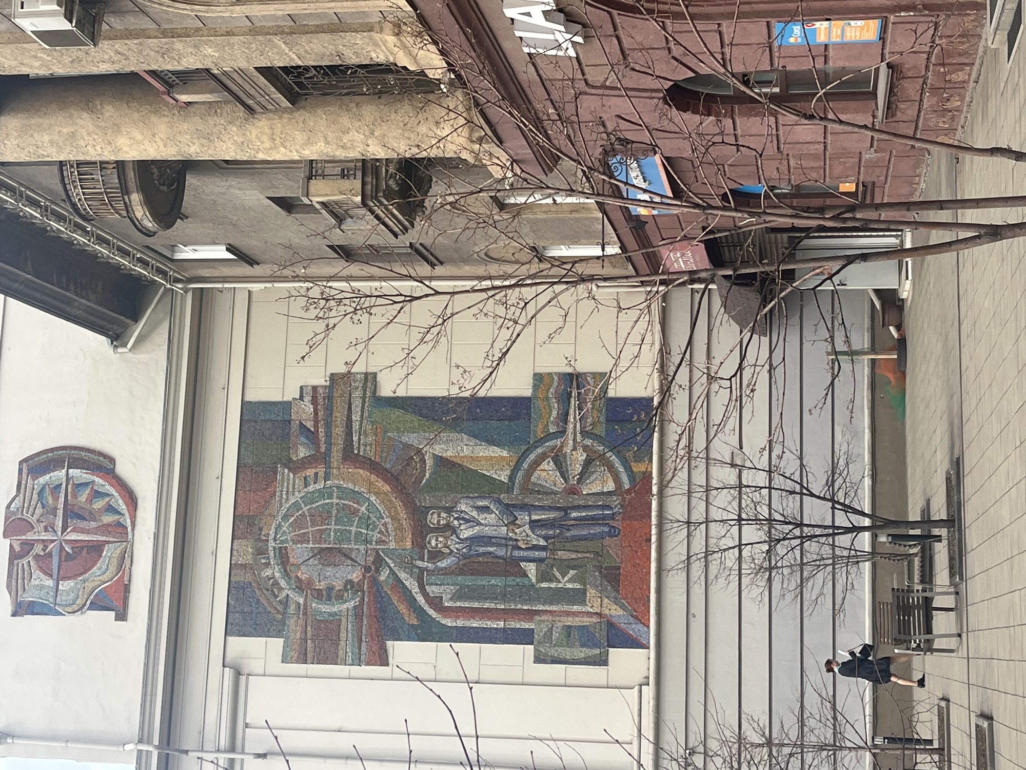 The image shows a building exterior in an urban environment, likely a city center. A large mosaic mural is visible on the building's facade, featuring abstract geometric designs and human figures, suggesting a mid-20th century style. A smaller mosaic is also present. Bare tree branches obscure parts of the view, indicating a colder season. The architecture includes elements of older, possibly historical buildings in the background, along with more modern structures. A person is walking along the wall, passing the mural. Street furniture, such as benches, is positioned in front of the building.