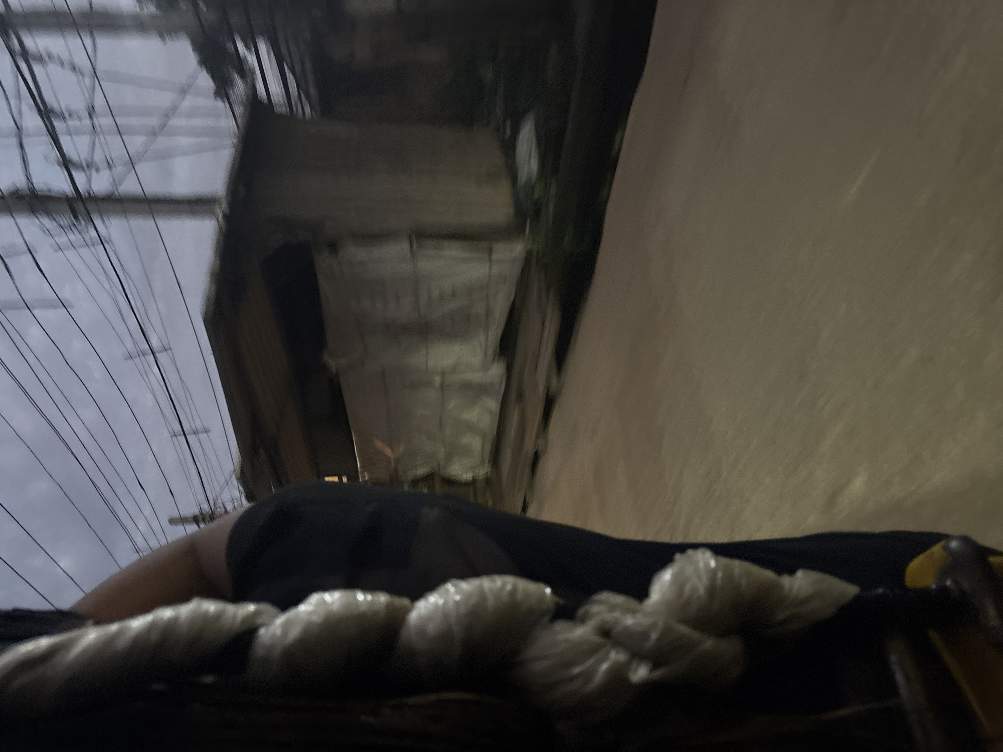 The image depicts a dimly lit street scene, likely in the city of Onitsha, Nigeria, as suggested by the context. The foreground is dominated by the side of a person wearing dark clothing, with what appears to be a string of wrapped objects hanging from their side. These objects are pale and roughly spherical, covered in a translucent material, possibly plastic. The person's arm is visible, resting on what could be part of a vehicle or structure.
In the background, a low-lying building with a corrugated metal roof is visible, partially obscured by white sheeting or tarpaulin stretched across its front. The building's structure appears rudimentary. Above the building, a dense network of power lines crisscrosses the sky, supported by utility poles, indicative of an urban environment with visible electrical infrastructure. The sky is overcast, suggesting either early morning, late evening, or generally cloudy weather, contributing to the overall low light conditions of the scene. The ground surface appears to be a paved road, reflecting the ambient light and showing signs of wear. The image's motion blur suggests it was taken while in transit, possibly from a moving vehicle.
