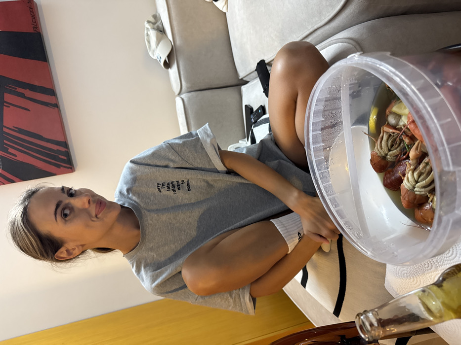 A fair-skinned woman sits sideways on a light-colored sofa. She is wearing a gray t-shirt with a black inscription, white socks, and has blonde hair. A clear bucket of crawfish is visible, with a bottle of beer resting near the sofa's edge. A modern artwork is displayed on the wall behind her, featuring red and black abstract designs. The sofa has light gray cushions and a small remote control is visible on the sofa. A portion of a dark wooden table appears in the lower right corner.