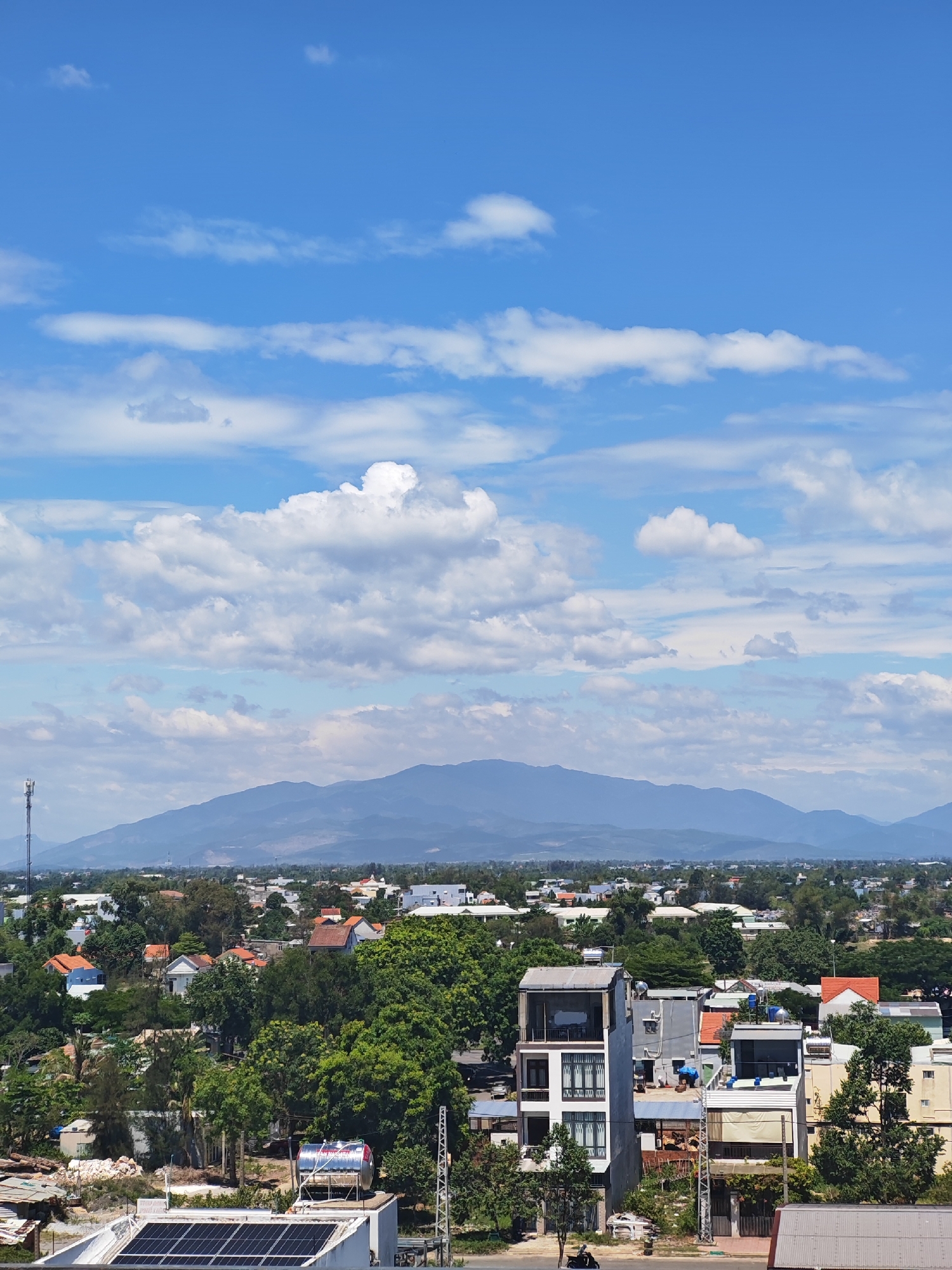 The image displays a panoramic view of a city, likely in Đà Nẵng, Vietnam, under a bright blue sky dotted with fluffy white clouds. In the foreground, a multi-story building with a modern, somewhat utilitarian design is visible, featuring balconies and dark-paned windows. Solar panels are installed on the rooftop of a lower structure to the left, suggesting a focus on renewable energy. The surrounding area is densely populated with various residential buildings, many with terracotta roofs, interspersed with lush green trees and vegetation. In the distance, a range of mountains forms a hazy blue backdrop against the horizon. A solitary cell tower stands on the left side of the frame, indicating telecommunications infrastructure. The scene appears to be a typical urban landscape in a tropical or subtropical region, with a mix of traditional and modern architecture. No people are explicitly visible in the image, though the presence of vehicles suggests ongoing activity. The overall impression is one of a developing urban center with a natural landscape in the background.