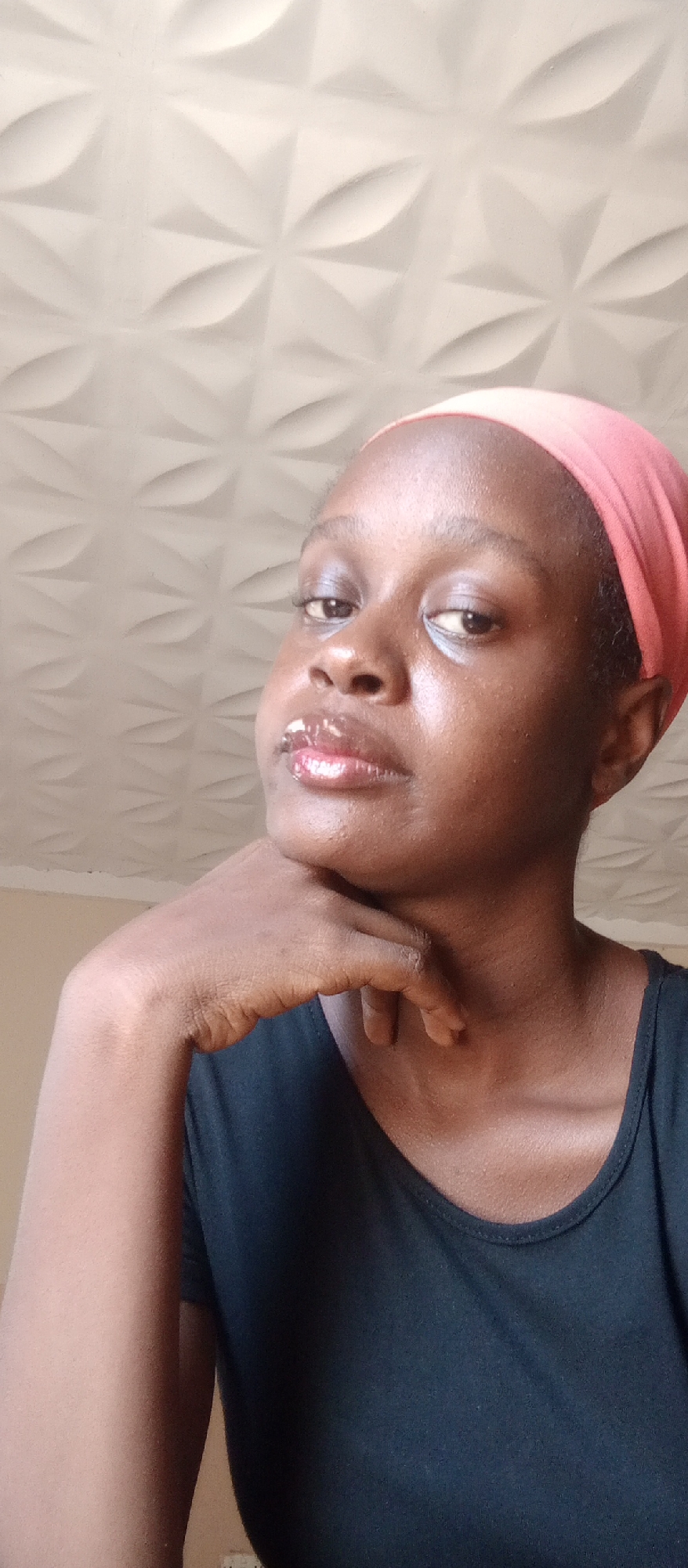 Akwanga, Nigeria: A medium, close-up shot depicts a dark-skinned woman with a pink headwrap, resting her chin on her left hand. She wears a black top. The background features a beige-colored wall and a patterned white ceiling.