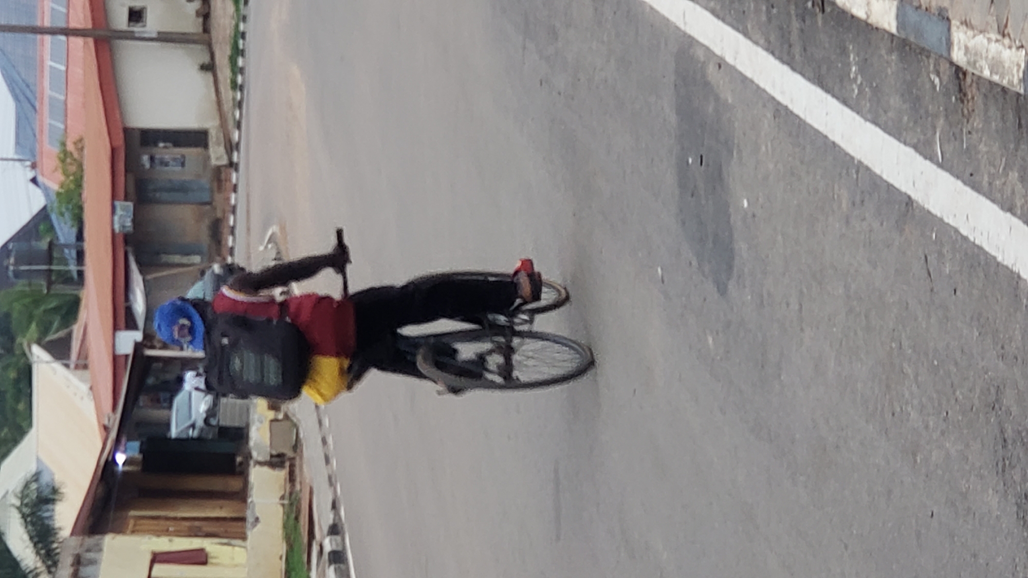 The image, oriented vertically and rotated 90 degrees clockwise, displays a person riding a bicycle on a paved road. The central figure, a cyclist with dark skin, is positioned in the middle-left of the frame. They are wearing a blue head covering or hat, a red shirt with a white stripe, black trousers, and red shoes. A black backpack is strapped to their back, and a bright yellow object, possibly another bag, is visible below the backpack, attached to either the person or the bicycle. The bicycle itself is dark-colored.
The road surface is asphalt, featuring a clear white line that demarcates the edge or a lane, running diagonally from the top-right to the bottom-left of the image. Along the side of the road, which appears at the top of the rotated image, a collection of buildings is visible. One building stands out with a reddish-brown roof, featuring what appear to be solar panels. Other structures showcase varied architectural elements, including painted facades and sections of exposed brick. Scattered green vegetation and a utility pole are also discernible among the buildings. The scene is illuminated by bright daylight, suggesting clear weather. No discernible text is visible anywhere in the image. The setting appears to be an urban or semi-urban area, consistent with the specified location of Narayi, Nigeria.