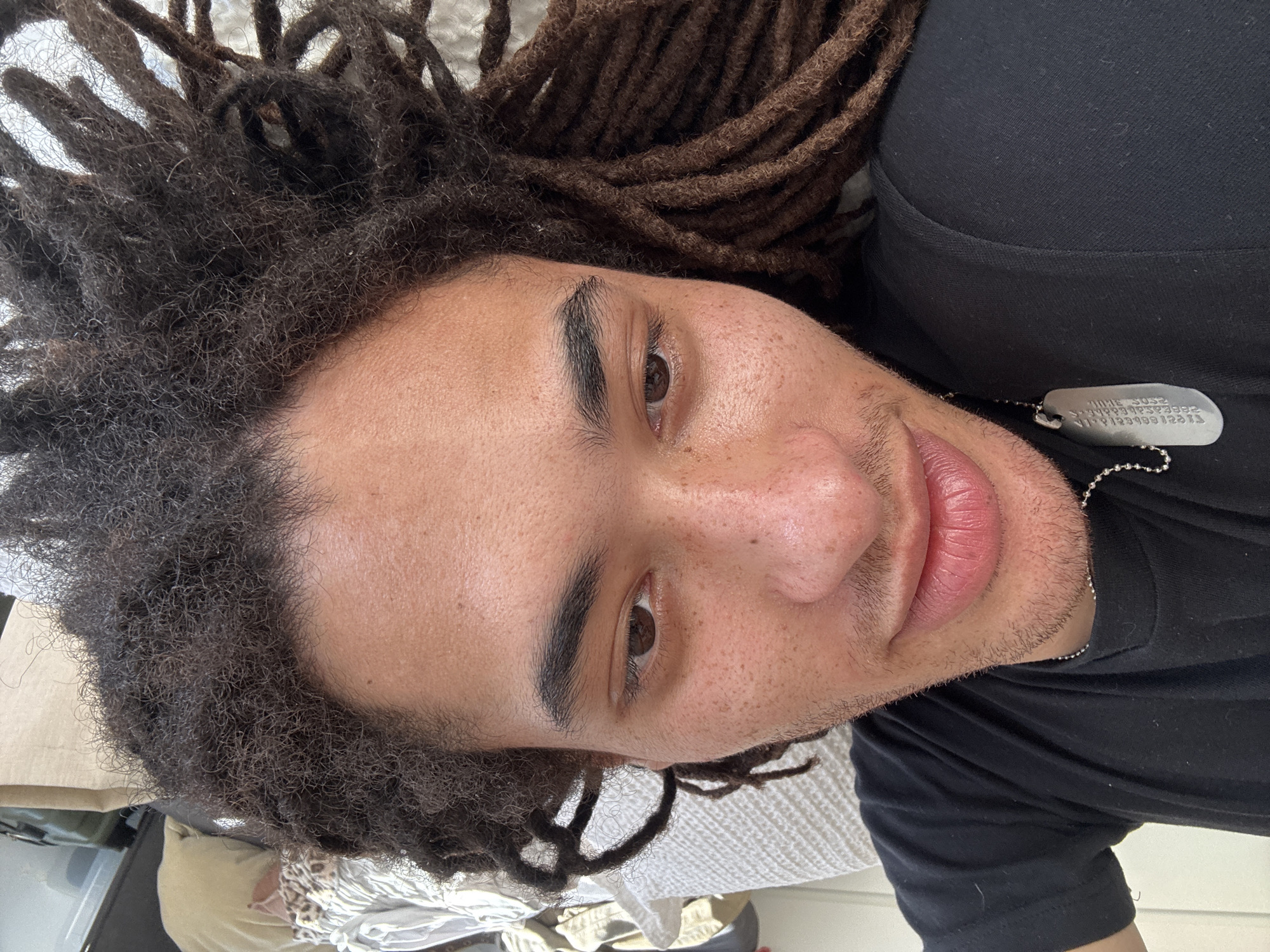 A close-up, tilted portrait features a young man with brown eyes and light brown skin. He has locs; some are dark brown and coiled, while others are looser and more textured near his hairline. He is wearing a black shirt and a silver dog tag necklace. His lips are full, and he has a slight smile. Freckles are visible on his nose and cheeks. The background appears to be a bed with white bedding. The location cannot be determined from the image provided.