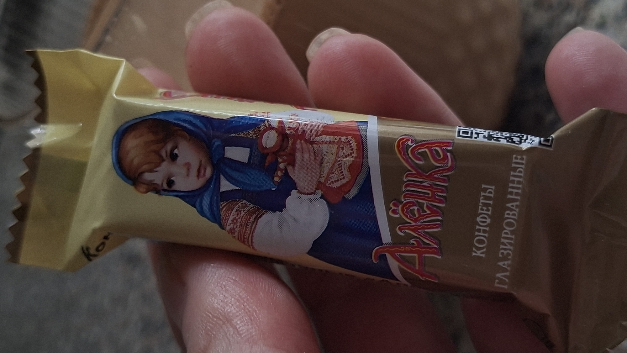 A hand holds a wrapped candy bar, with a yellow front and brown back, featuring a depiction of a young girl in traditional Russian dress. The girl has red hair and is wearing a blue headscarf and a blue dress with embroidered details. She is holding a small doll. Below the image of the girl, the word "Алёнка" is written in stylized pink and gold lettering. Further down, text in white reads "КОНФЕТЫ" and "ГЛАЗИРОВАННЫЕ", indicating "Candies" and "Glazed" in Russian. A QR code is visible on the right side of the wrapper. The background is blurred, suggesting it's an indoor setting, possibly a home. There is no visible indication of the city of Myadzyel, Belarus, in the image itself.
