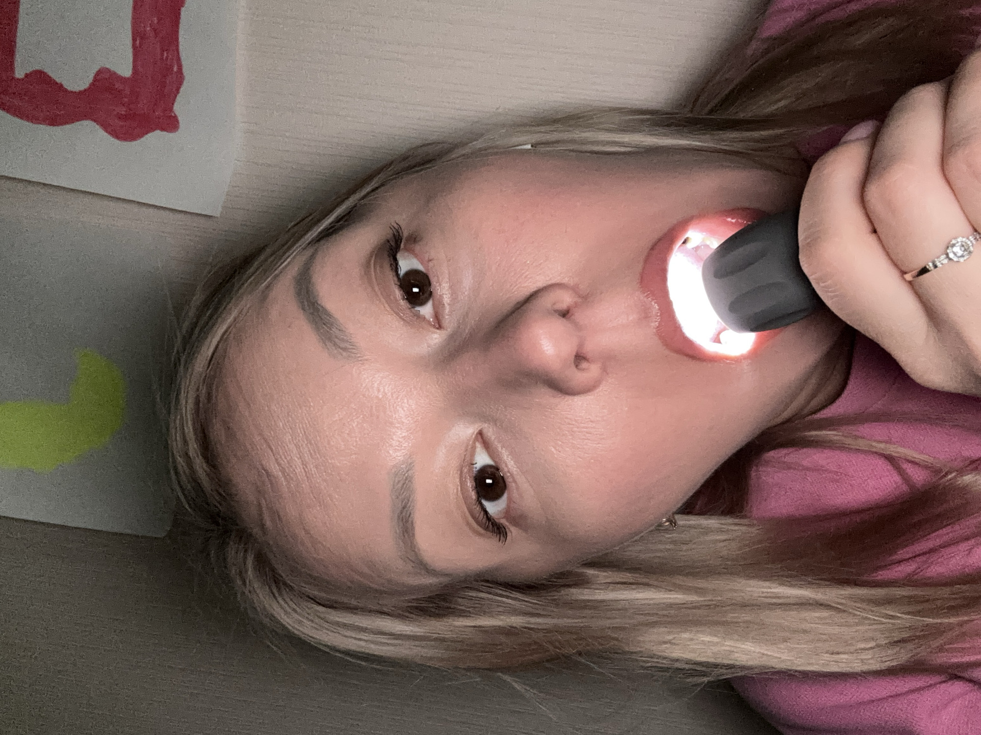 A woman with blonde hair is holding a black flashlight in her mouth. She is wearing a pink shirt and a ring on her finger. There is some artwork in the background. The location is not identifiable as Izhevsk, Russia.