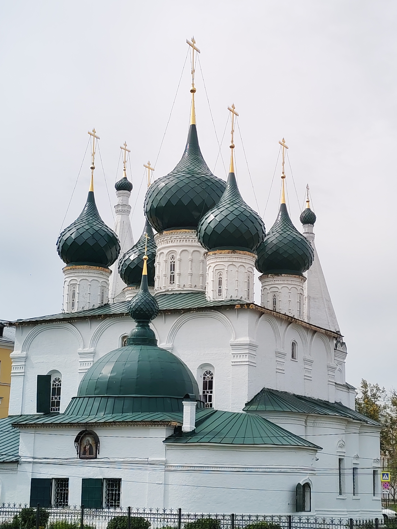 The image shows the Assumption Cathedral in the city of Zhukovo, Russia, under an overcast sky. The cathedral is a white building with multiple green, onion-shaped domes topped with golden crosses. The domes vary in size and are arranged in a clustered pattern. White decorative elements and arched windows are visible on the exterior walls. In the foreground, there is a black metal fence and some greenery. To the left, a yellow building is partially visible, and to the right, trees and a street sign with a pedestrian crossing symbol can be seen. The lower section of the cathedral features a large green dome and several windows with green shutters. An icon depicting a religious figure is mounted on the white wall.
