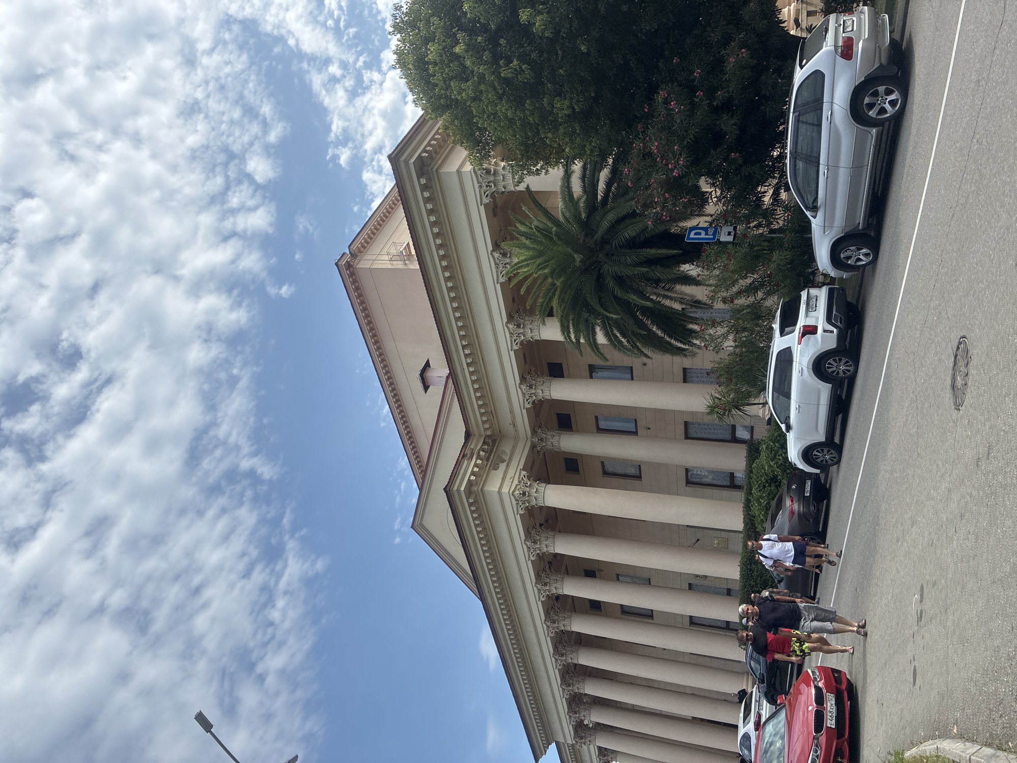 This image captures a sunny day in Sochi, Russia, in front of a grand, colonnaded building, likely a public institution or theater, given its neoclassical architecture. Several people are walking on the sidewalk and street. A red BMW sedan is parked on the left, and a silver BMW SUV is parked on the right. A white SUV is also present. The building features a large number of columns and ornate detailing, with a palm tree and lush greenery to the right. The sky is blue with scattered white clouds. The overall atmosphere appears to be relaxed, with people dressed in casual summer attire.
