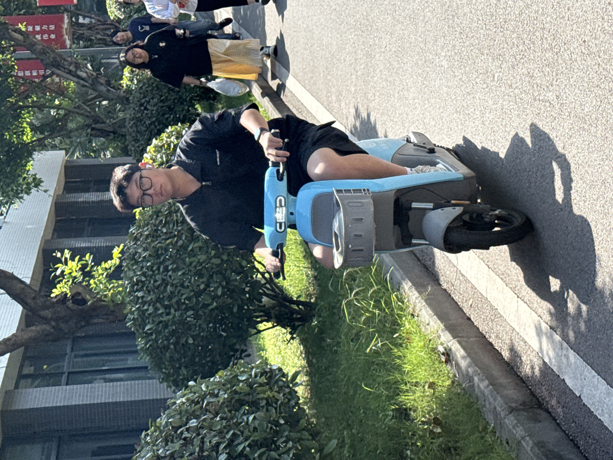 This brightly lit outdoor photograph captures a young man riding a light blue electric scooter on a sunny day in what appears to be an urban or campus setting in Jiangjin, China. The man, positioned prominently in the foreground, wears eyeglasses, a black short-sleeved top, and black shorts, with a watch visible on his left wrist as he holds the scooter's handlebars. His expression is neutral.
The light blue electric scooter features a modern design. Strong sunlight creates distinct shadows of the scooter and nearby elements on the gray asphalt ground, indicating clear weather and either morning or afternoon. A white line is painted along the edge of the asphalt pathway. To the left, a concrete curb separates the path from a grassy area lined with lush green bushes and trees.
In the background, several other individuals are visible walking along a sidewalk or path. One woman in a black top and yellow skirt or carrying a yellow bag is seen, along with other people further back. A modern building with multiple windows is partially visible on the far left. A red sign with white and yellow Chinese characters, including "大学" (dàxué - university/college) and "新力" (xīnlì - new power/force), is partially obscured by trees in the mid-ground, further hinting at a university or urban location. The overall atmosphere is casual, reflecting everyday activity.