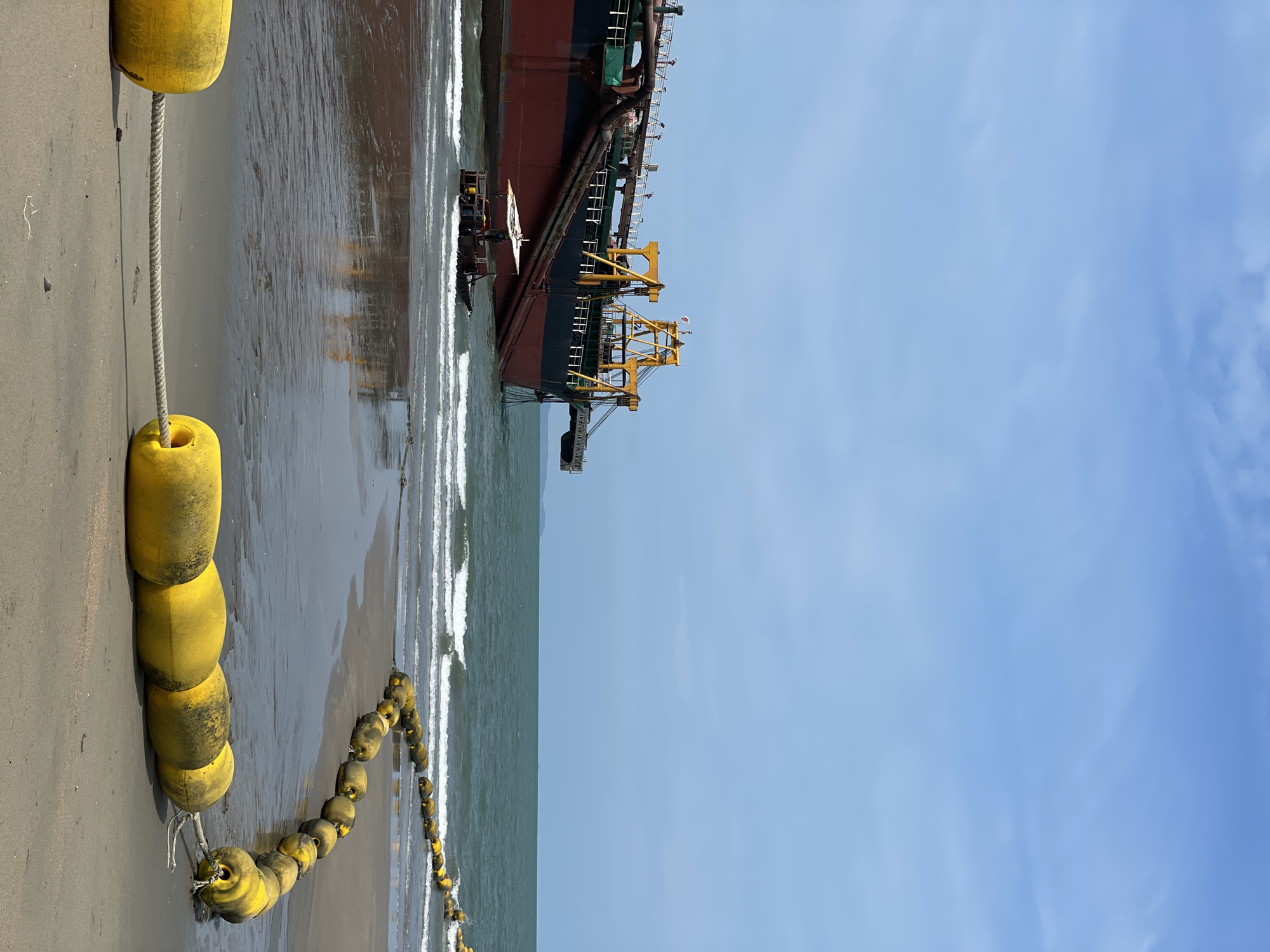 A large dredger ship is beached on a sandy shore in Pattaya City, Thailand. A chain of yellow buoys extends along the shore, partially submerged in the water. The sky is clear and blue, with some cloud cover. Waves break on the beach next to the ship.