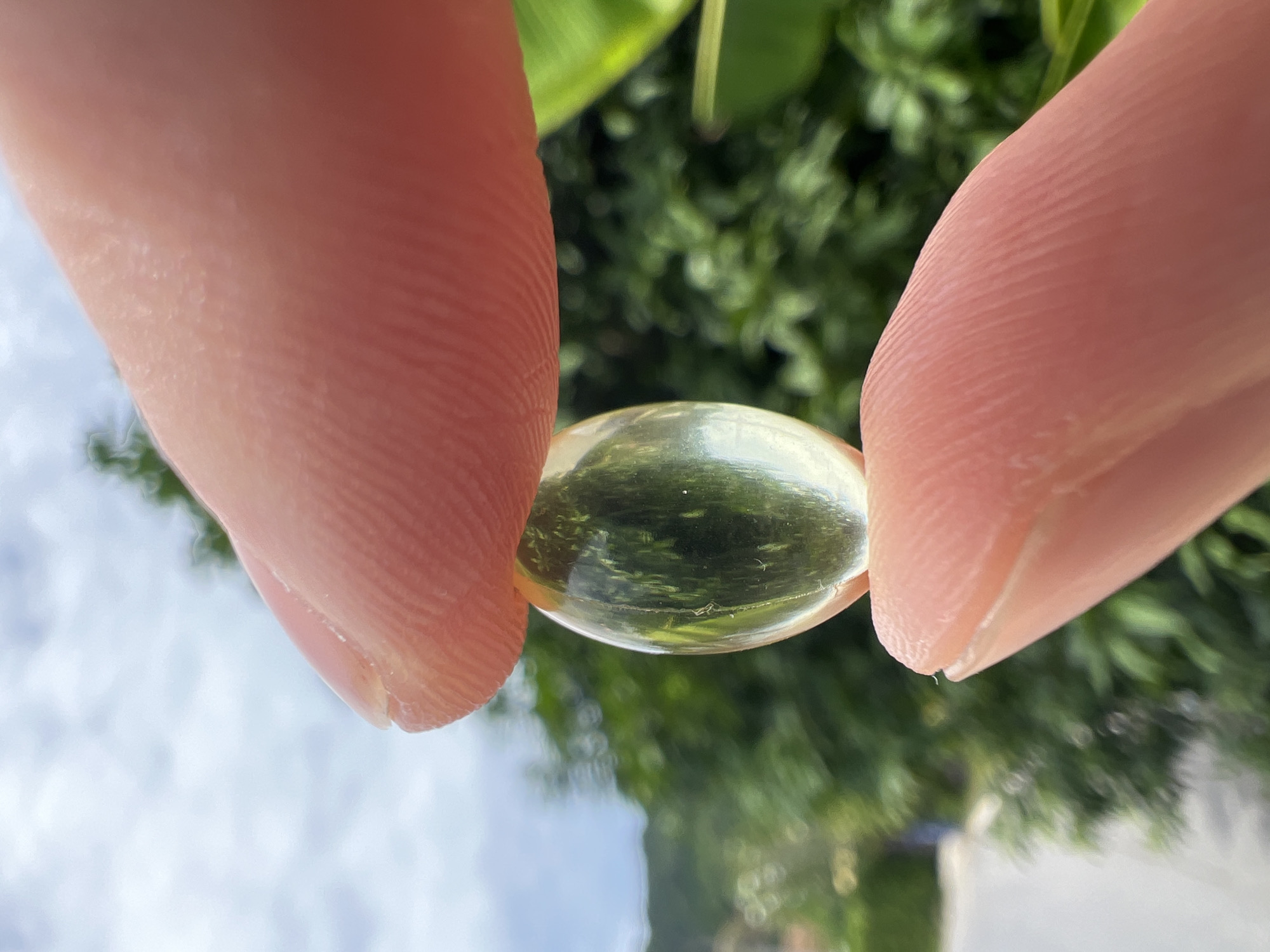A close-up, vertical, outdoor shot features two fingers holding a translucent, oval-shaped capsule. The capsule is yellow and appears to contain a liquid. The fingers, with visible fingerprints, are positioned to grip the capsule, creating a slight distortion in its shape. The background is blurred, showing lush green foliage and a bright sky with scattered clouds, suggesting a tropical environment. A distant road and hint of buildings are visible in the lower left portion of the image, indicating an urban or suburban setting within Choeng Thale, Thailand.
