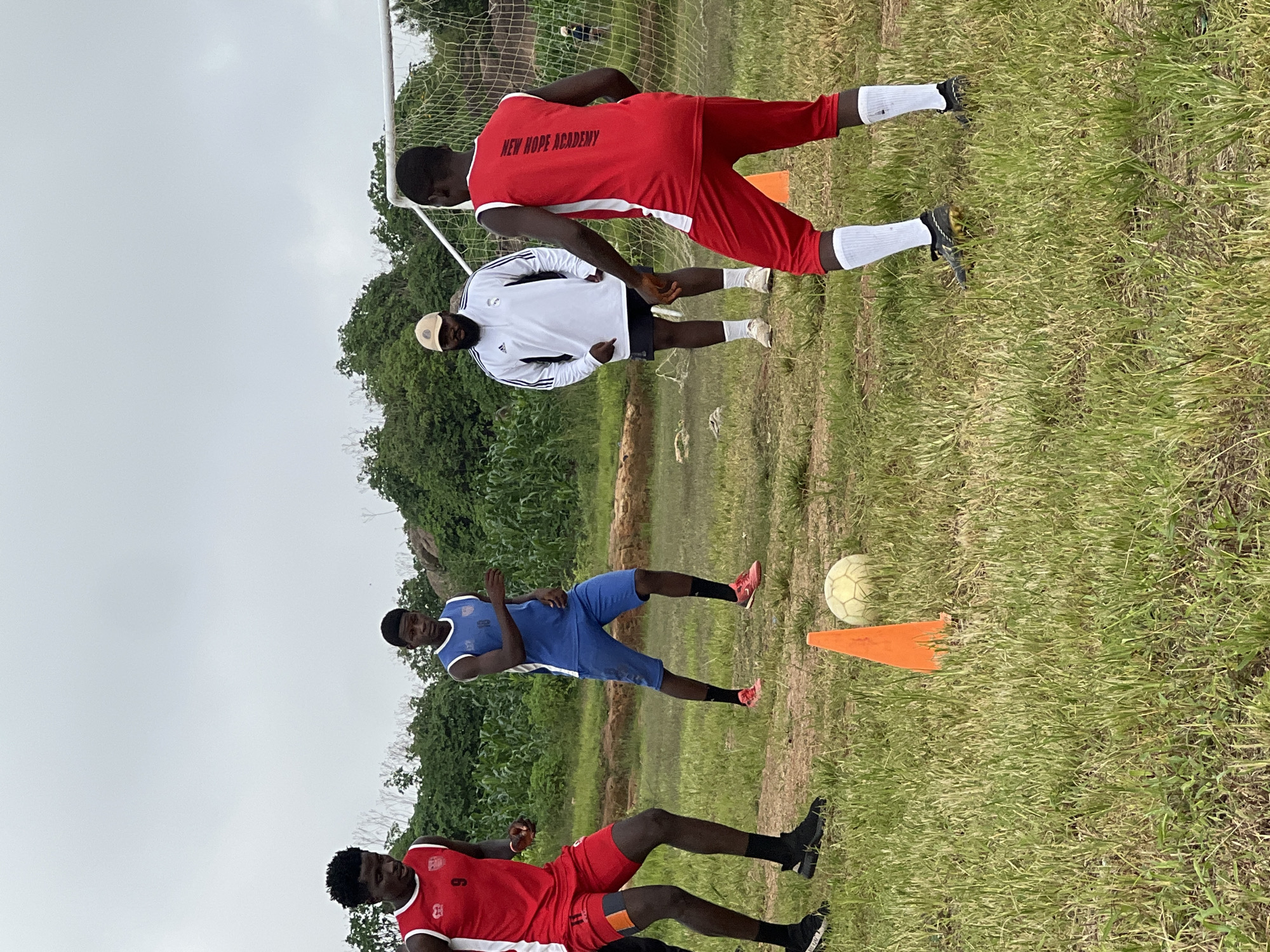 In Jos, Nigeria, four young men are engaged in a soccer practice session on an outdoor field. Two of the men, wearing red jerseys and shorts, are positioned on the left and right sides of the frame, respectively. The man on the left wears the number 9 on his jersey, while the man on the right has "NEW HOPE ACADEMY" written vertically on his back. Another young man in a blue jersey and shorts, with the number 18 on his jersey, stands in the center-left of the image. A fourth individual, an adult male wearing a white Adidas tracksuit and a beige cap, stands in the background, observing the players. The field is covered in green grass with some patches of dry earth. An orange cone and a soccer ball are visible in the foreground. In the background, a soccer goal with a net is set up, and lush green vegetation and some rocky outcrops are present. The sky is overcast, suggesting a cloudy day.