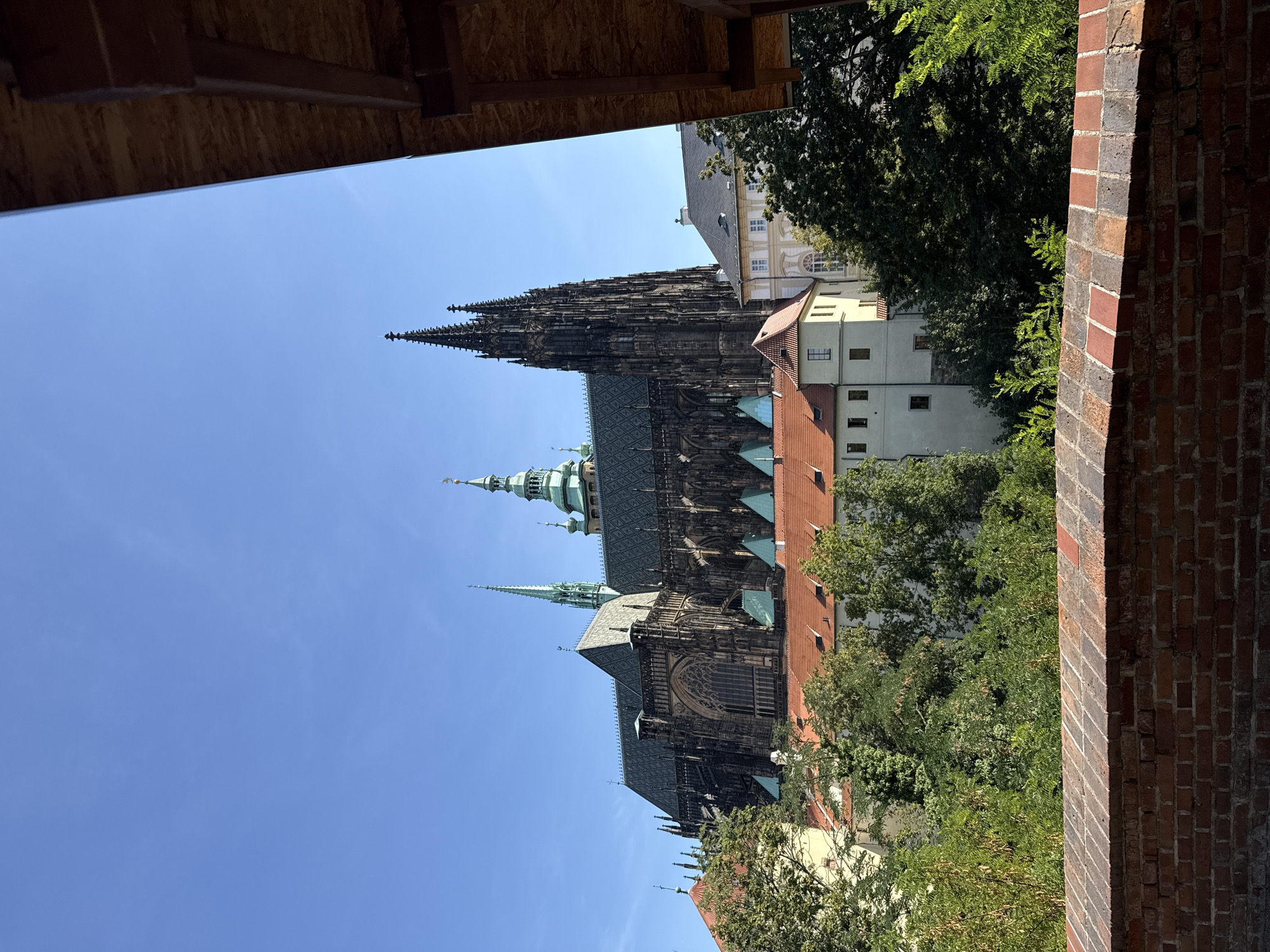 The image shows a view of St. Vitus Cathedral in Prague, Czechia, on a clear, sunny day. The imposing Gothic cathedral, with its dark stone facade, intricate spires, and arched windows, dominates the upper portion of the frame. In front of the cathedral are several buildings with red tile roofs and light-colored walls, interspersed with lush green trees. A brick wall runs along the bottom of the image, framing the scene. Part of a wooden structure is visible on the right side, obscuring a small portion of the sky. The sky is a vibrant, cloudless blue. There are no people or visible activity in the image.