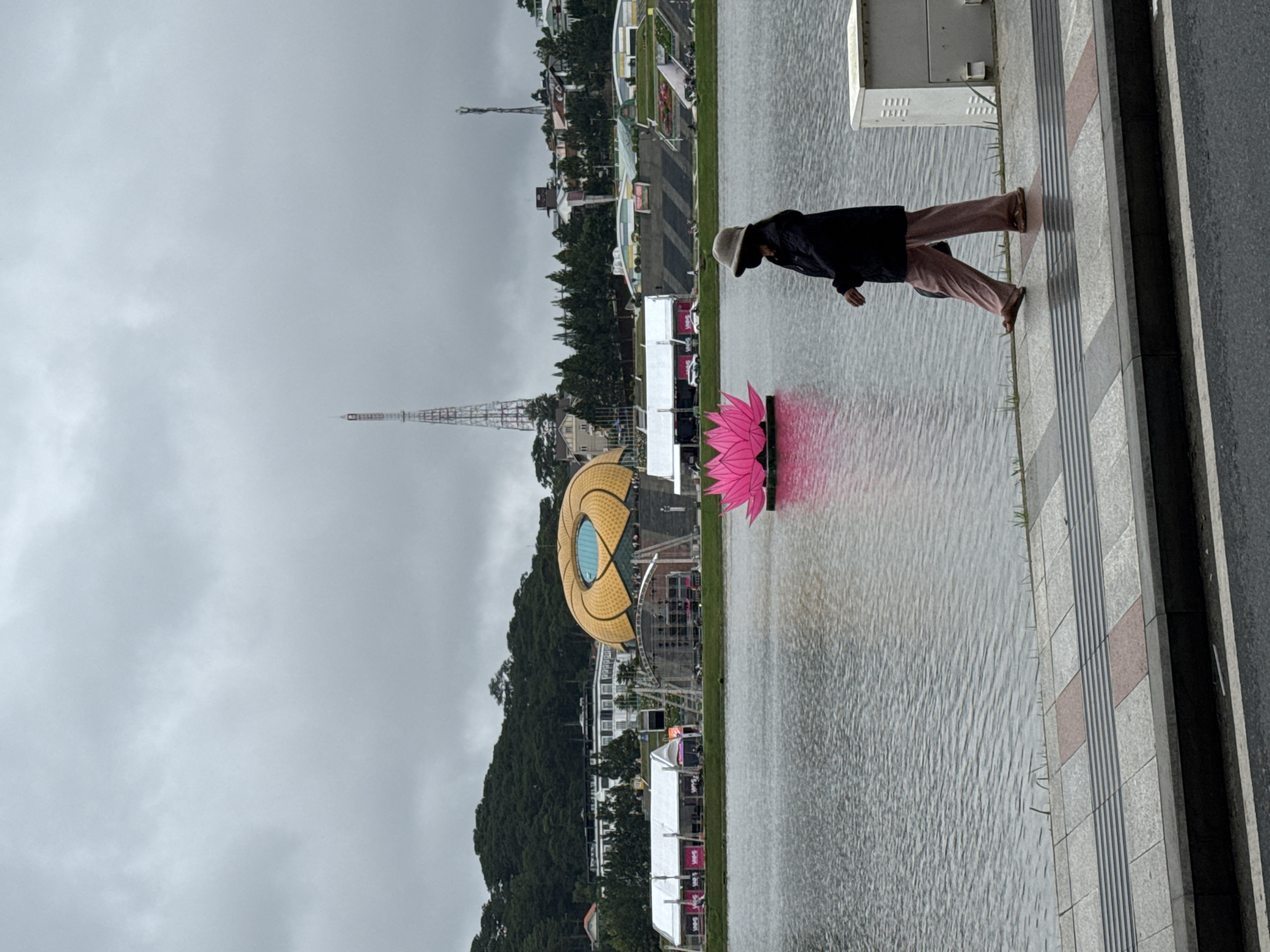 The image captures a scenic view of Xuan Huong Lake in Da Lat, Vietnam, characterized by an overcast sky. The scene is dominated by a large body of water on the left, reflecting the grey sky, and a paved pedestrian walkway on the right.
A lone person stands on the walkway in the mid-ground on the right, seen from the back and side. They wear a dark jacket, light-colored pants, and a hat, seemingly observing the lake. Adjacent to the person on the walkway is a grey utility box or cabinet.
Prominently featured in the water, closer to the foreground, is a large, illuminated pink lotus-shaped structure, likely an art installation or decoration for an event. Further across the water, on the distant shore, a distinct golden, eye-shaped building with a blue circular center, characteristic of the Da Lat Lam Vien Square, stands out. A tall, lattice-style transmission tower or radio mast is visible in the far background.
Along the lake's edge, white tent-like structures, possibly event booths, are set up. The distant shore is lined with lush green trees and various buildings. The sky is uniformly grey and cloudy, indicating an overcast day. The water surface shows subtle ripples. The paved walkway features tactile paving strips. While some faint text is present on the white tents, it is too small and indistinct to be read clearly. The scene suggests a public gathering space or a city festival setting.