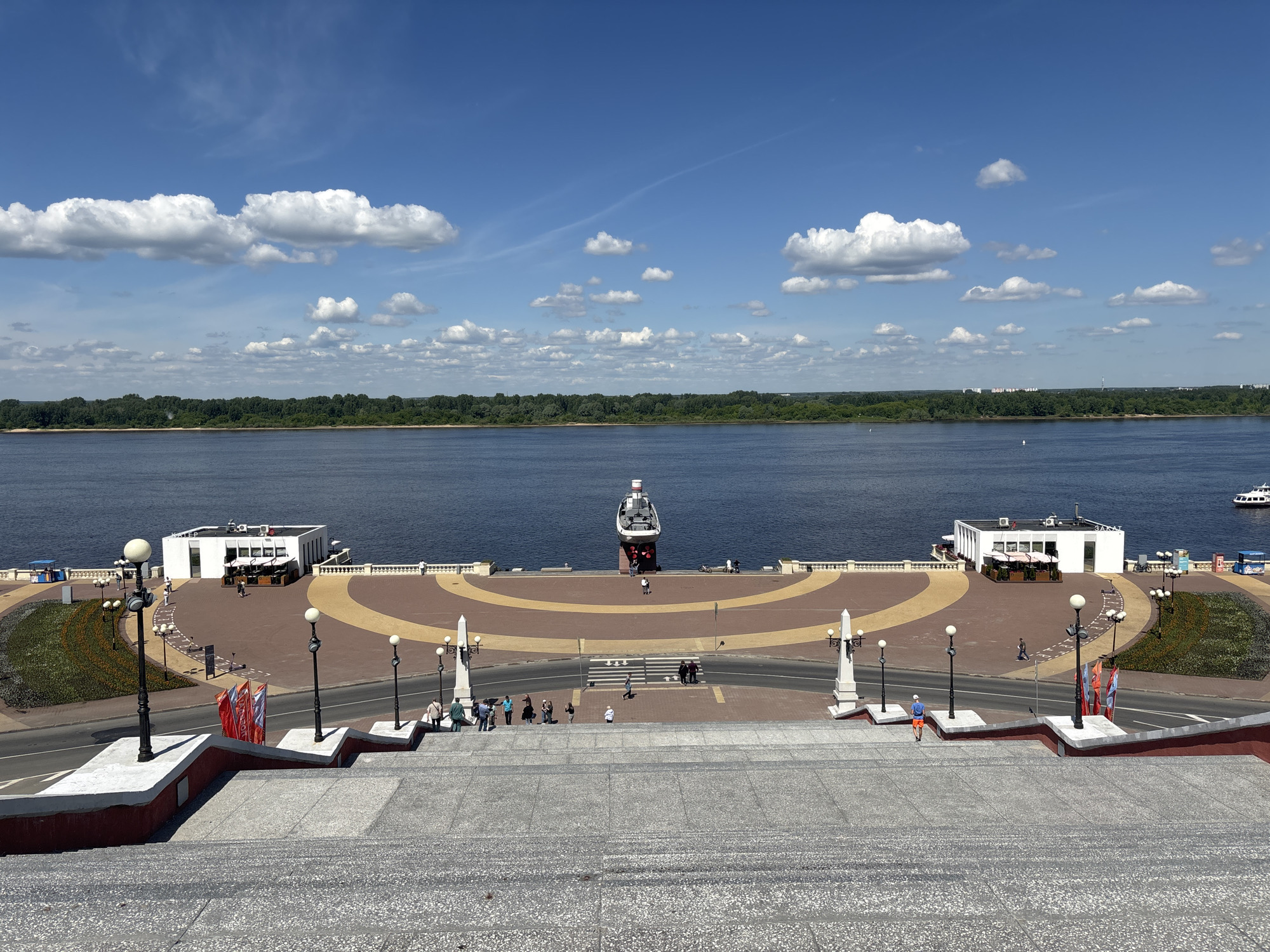 The image presents a high-angle view of a bustling river embankment in Nizhny Novgorod, Russia, seen from the vantage point of a wide, grey-speckled staircase. The scene is bathed in bright daylight under a clear blue sky adorned with scattered white cumulus clouds.
In the foreground, the broad staircase, featuring red and white borders, descends towards a paved road with a crosswalk. Beyond the road lies a spacious, semicircular promenade, paved with light and darker brown patterns. This embankment is furnished with numerous ornate lampposts and features landscaped areas with greenery.
Centrally located at the river's edge of the promenade is a prominent grey and white memorial ship, seemingly mounted on land as a monument. Two modern, white single-story buildings with large windows, likely cafes or restaurants, flank the ship. On the building to the right, the Cyrillic text "ЗАКАT" (ZAKAT) is visible. White balustrades line the entire riverfront edge of the promenade.
Numerous people are dispersed across the embankment and road, strolling individually or in small groups, suggesting a leisurely atmosphere. One person in orange shorts and a blue shirt is seen descending the staircase.
The expansive, dark blue river forms the middle ground, stretching horizontally across the frame. On the far bank, a dense line of green trees creates a natural backdrop, with faint hints of distant urban structures. A white pleasure boat is partially visible on the far right of the river, and small white buoys dot the water. The image captures a lively and scenic riverfront public space.