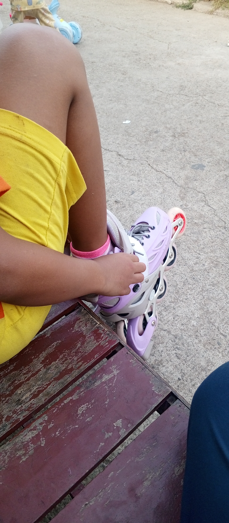 In a close-up shot, a person is sitting on a weathered red wooden bench, with their legs crossed. The person is wearing a bright yellow t-shirt and pink socks with white stripes. On their left foot, they are wearing a light purple inline skate with grey laces and pink wheels. The person's hand is resting on the skate. The background consists of a concrete surface and some distant green foliage. The lighting suggests it is daytime. The scene appears to be outdoors, possibly in a park or recreational area.