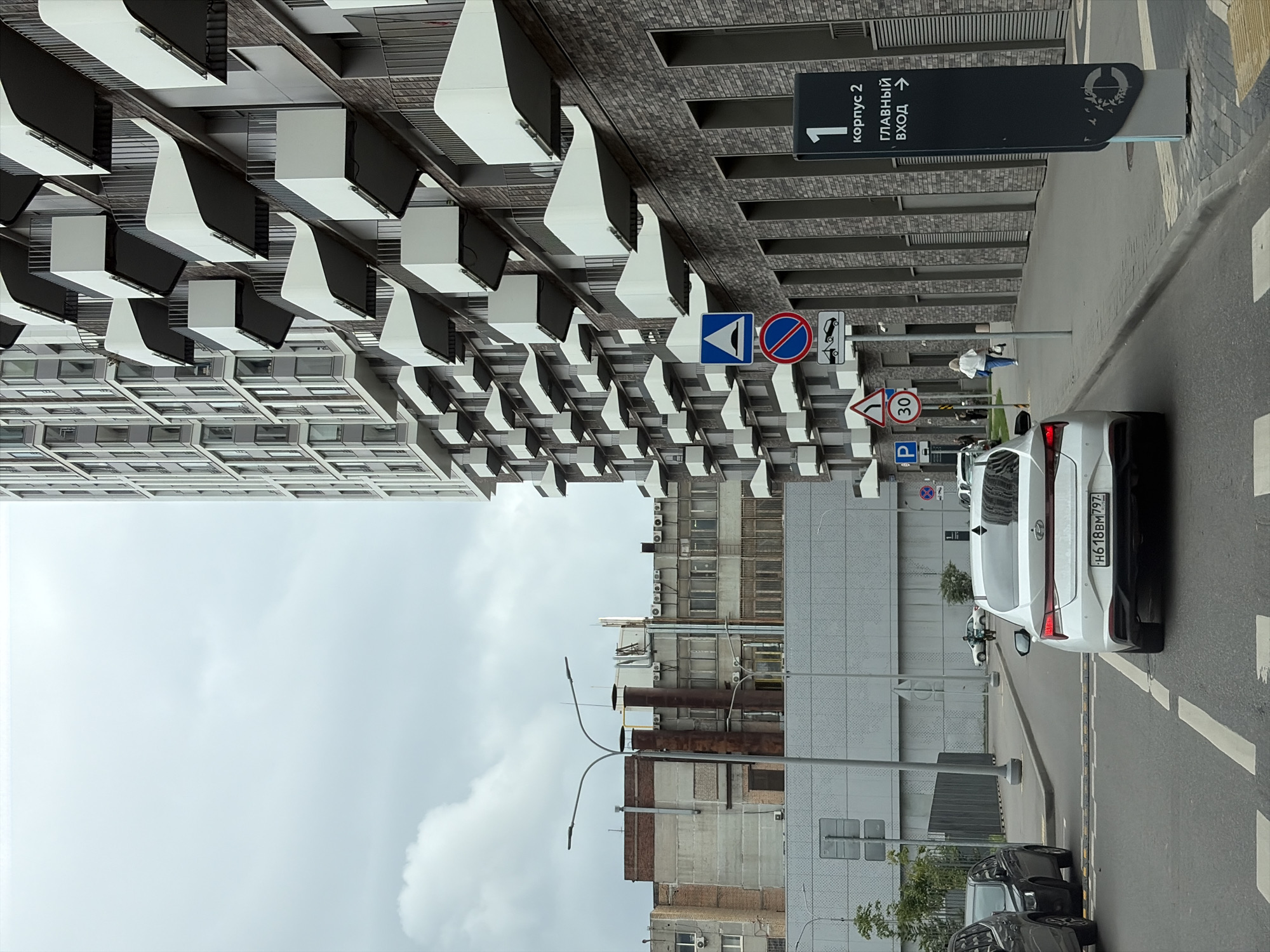 This image captures a street scene in Moscow, Russia, dominated by a modern, multi-story apartment building with numerous cantilevered balconies. To the left of the building, older, industrial-style structures are visible under a cloudy sky. The foreground shows a street with several cars, including a white sedan in the center with a Russian license plate H618BM797. A woman in a white shirt and blue jeans walks on the sidewalk in the middle ground, passing a series of traffic signs, including a speed limit sign indicating 30 km/h, a "no stopping" sign, and a sign with a towing truck icon. A tall, dark sign on the right side of the sidewalk displays Russian text, indicating "1 корпус 2" and "главный вход" (main entrance) with an arrow pointing to the right. The street is paved, with visible lane markings and a pedestrian crossing. The overall atmosphere is overcast and urban.