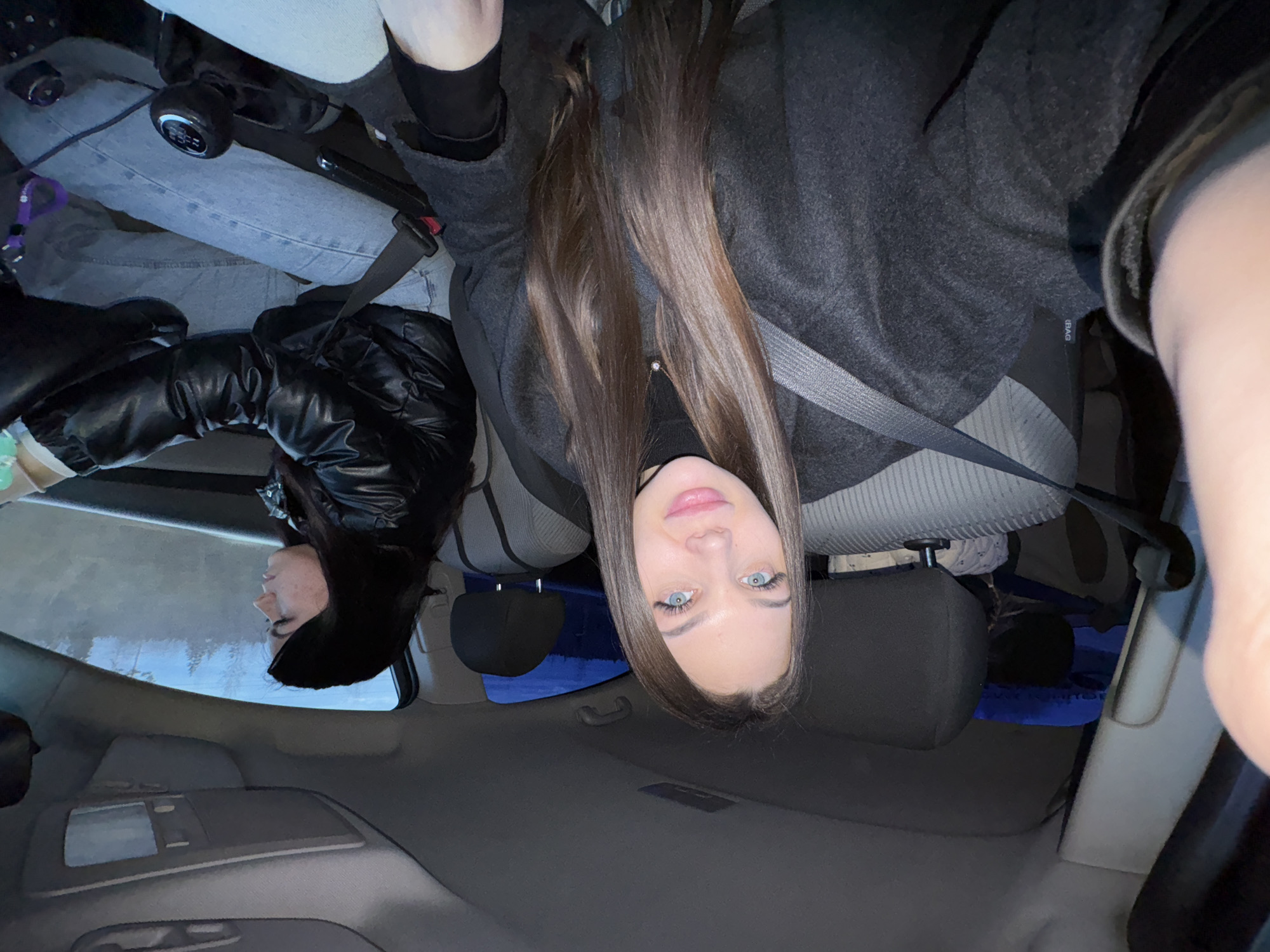 The image shows the interior of a car, oriented upside down. Two individuals are visible. One, likely the driver, is wearing a black leather jacket and a black hat. The other individual, presumably a passenger, has long brown hair, blue eyes, and is wearing a gray sweater. Both individuals are wearing seatbelts. The car's interior is gray, with light blue jeans visible on one of the individuals. The image location is not possible to determine from the provided picture.