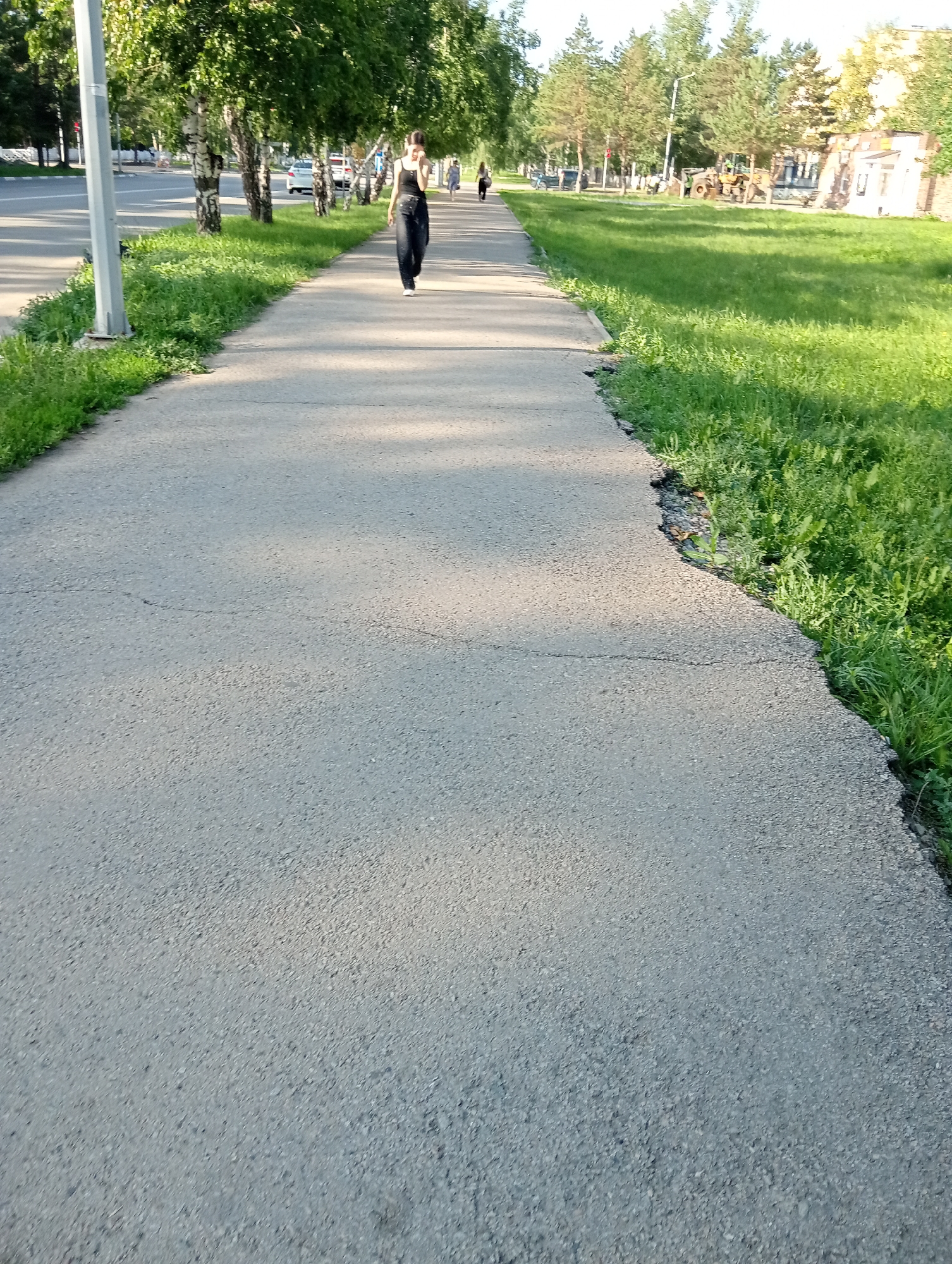 A person is walking away from the camera on a paved path. The path is bordered on the left by a street with cars and trees, and on the right by a grassy area. The person is wearing dark clothing and white shoes. In the background, there are more trees and buildings, including what appears to be construction equipment. The lighting suggests it is daytime, likely afternoon, with shadows cast across the path. The scene is an outdoor urban environment, possibly a park or a street in Kostanay, Kazakhstan.