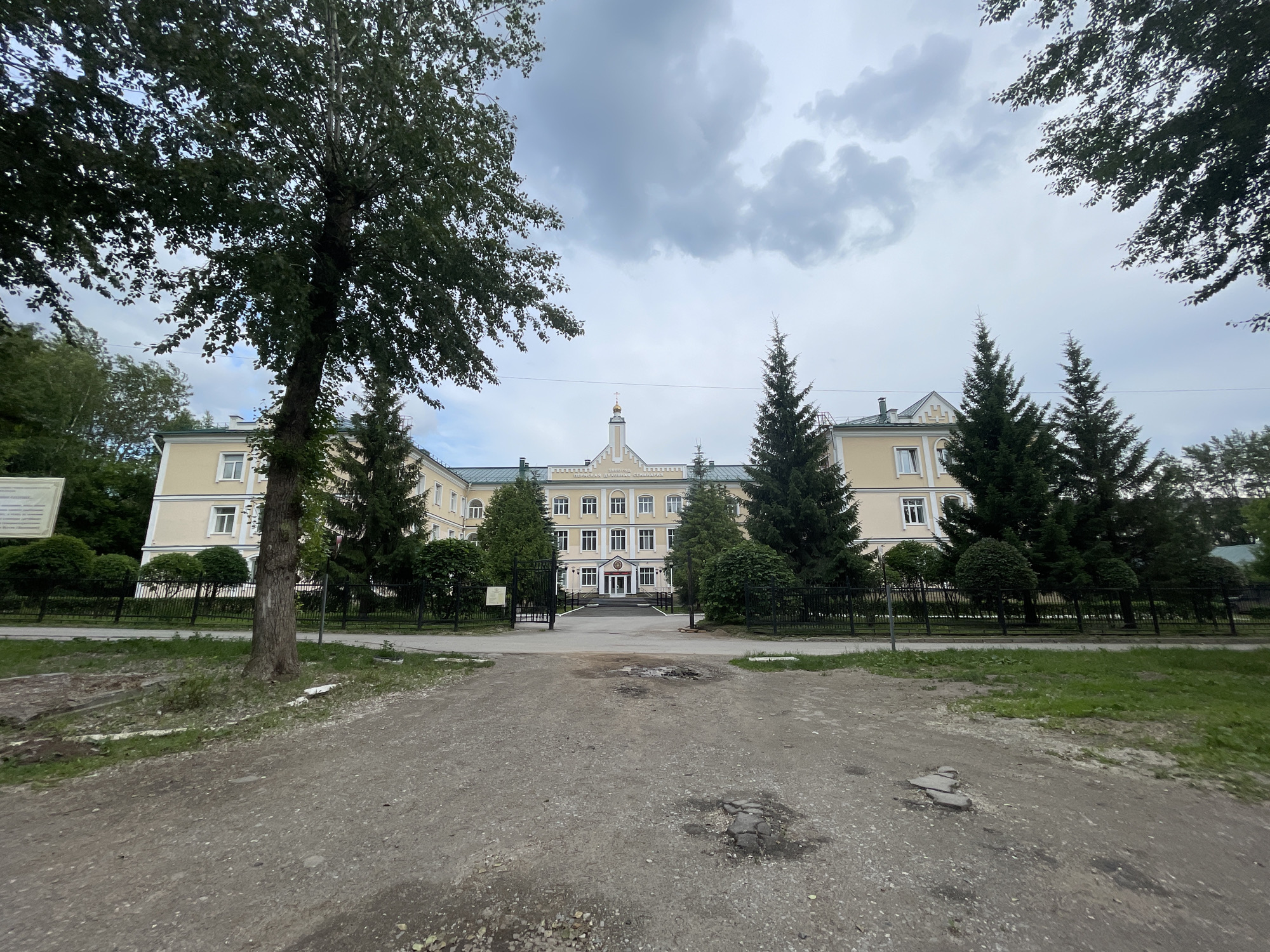 The image depicts the exterior of the Perm Theological School, located in Perm, Russia. The building is a long, light yellow structure with a green roof, featuring multiple windows and an arched entrance. A central tower with a golden dome and cross rises from the roof, topped by a spire. The building is surrounded by a black metal fence and lush greenery, including several tall evergreen trees and deciduous trees. The foreground shows a rough, gravelly path with some patchy grass and evidence of unrepaired road surface. The sky is overcast with grey clouds. There are no people visible in the image.